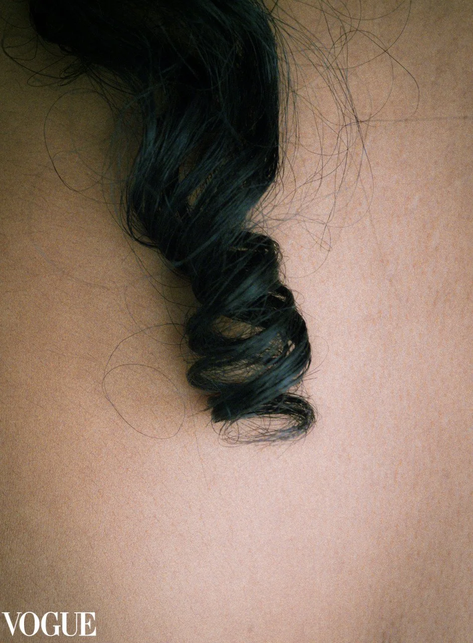 Close-up of a black curly hair strand on a person's skin, with the Vogue logo in the bottom left corner.