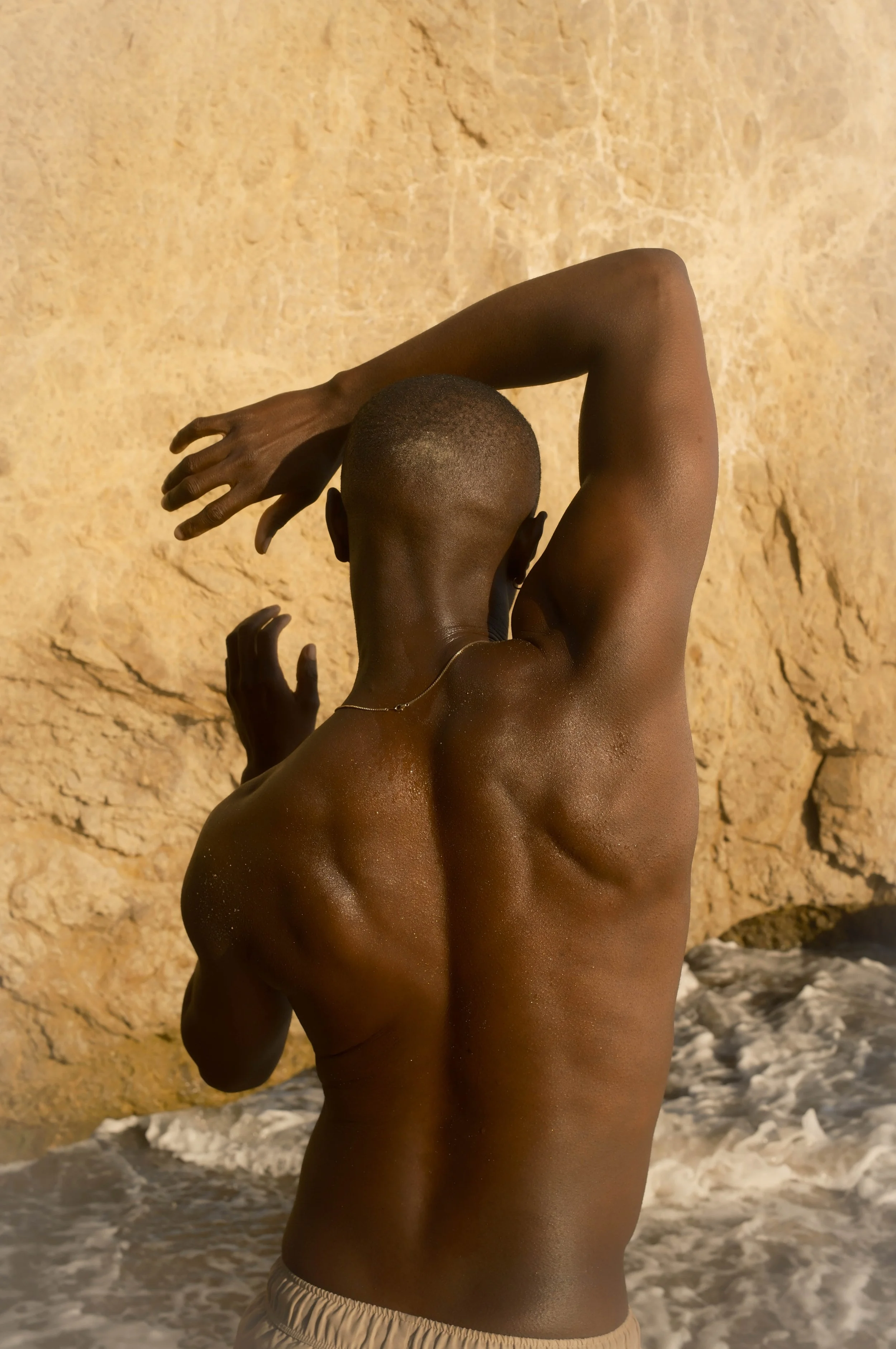 A person with dark skin and short hair standing in front of a rocky beach with waves.