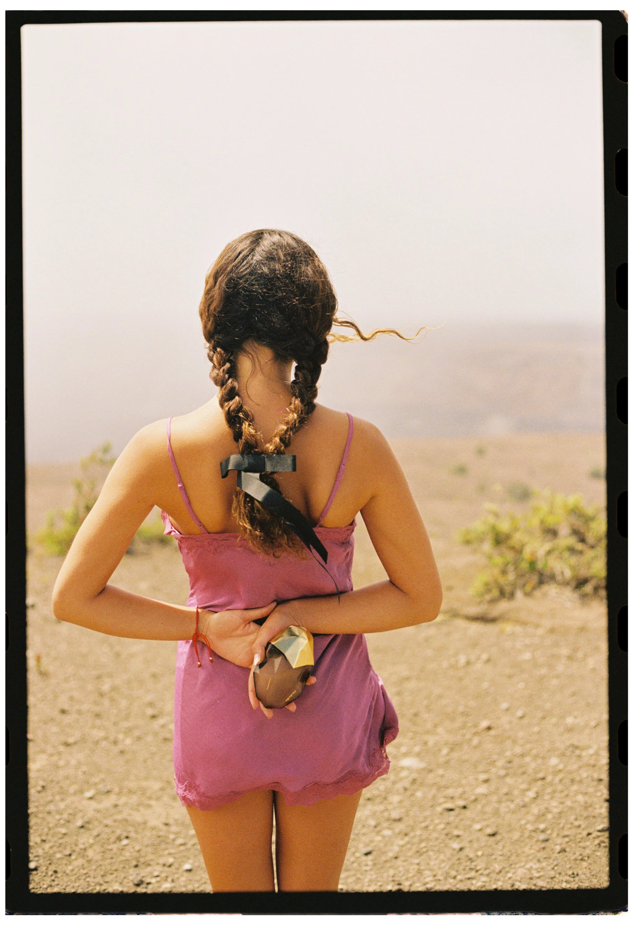 A woman with braided hair stands outdoors in a pink dress, holding a reflective object in her hands, with a beach and ocean in the background.