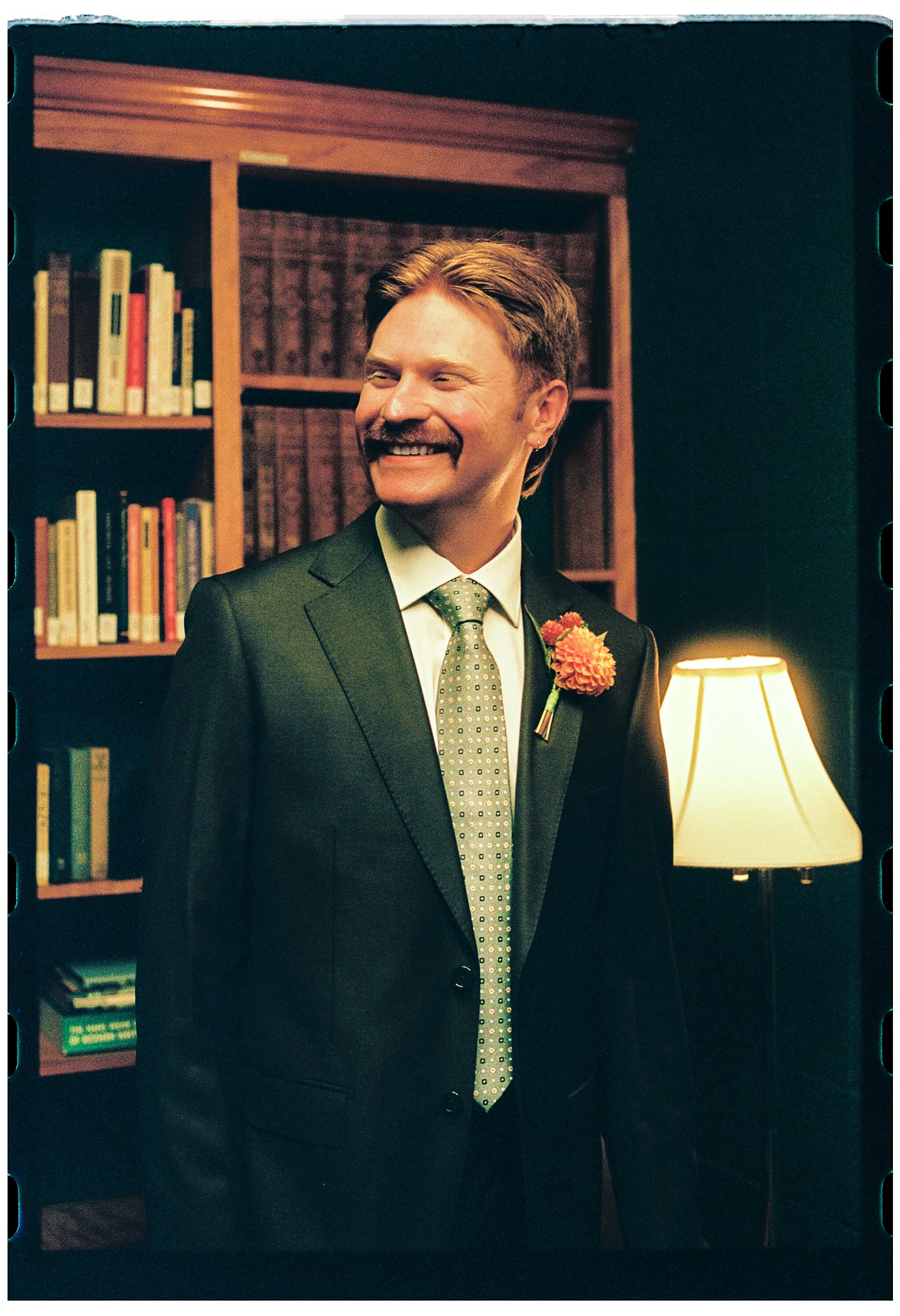 Smiling man in a suit standing in a room with bookshelves, a lamp, and warm lighting.