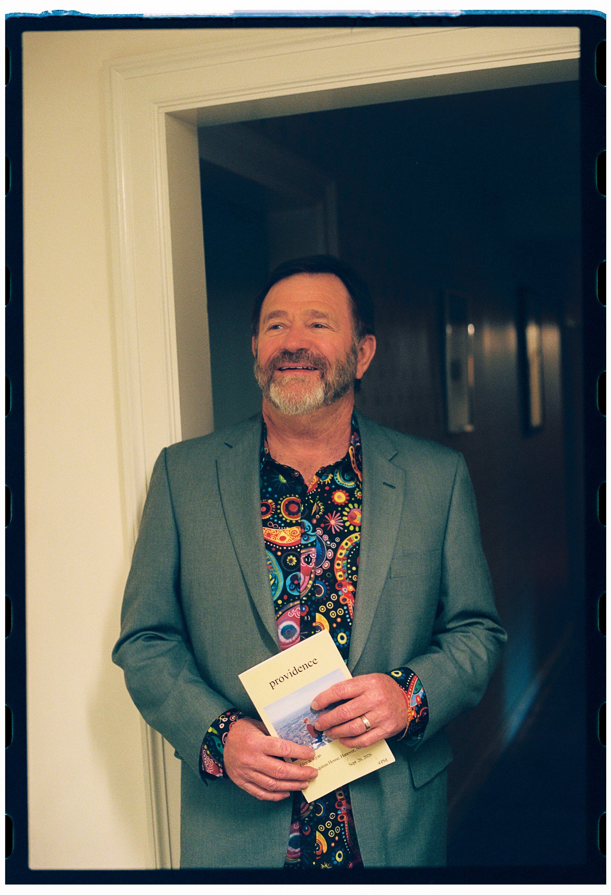 A smiling middle-aged man with a beard and mustache dressed in a gray suit jacket and a colorful paisley shirt, holding a brochure with the word 'Providence' on it, standing in front of a doorway in an indoor setting.