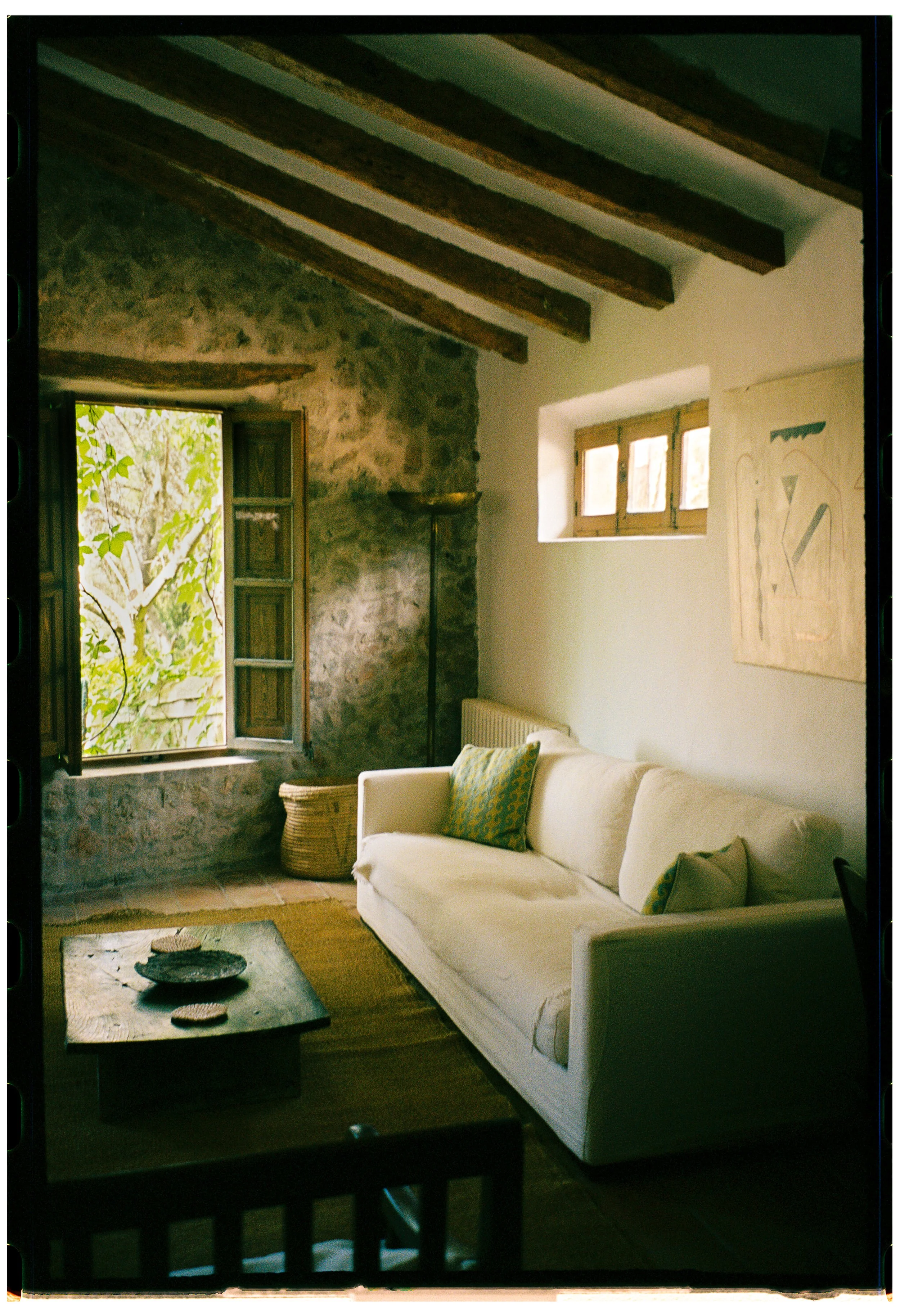 A cozy living room with a white sofa, decorative pillows, a stone wall with an open window, wooden beams on the ceiling, and a small window with natural light, decorated with art and greenery outside.
