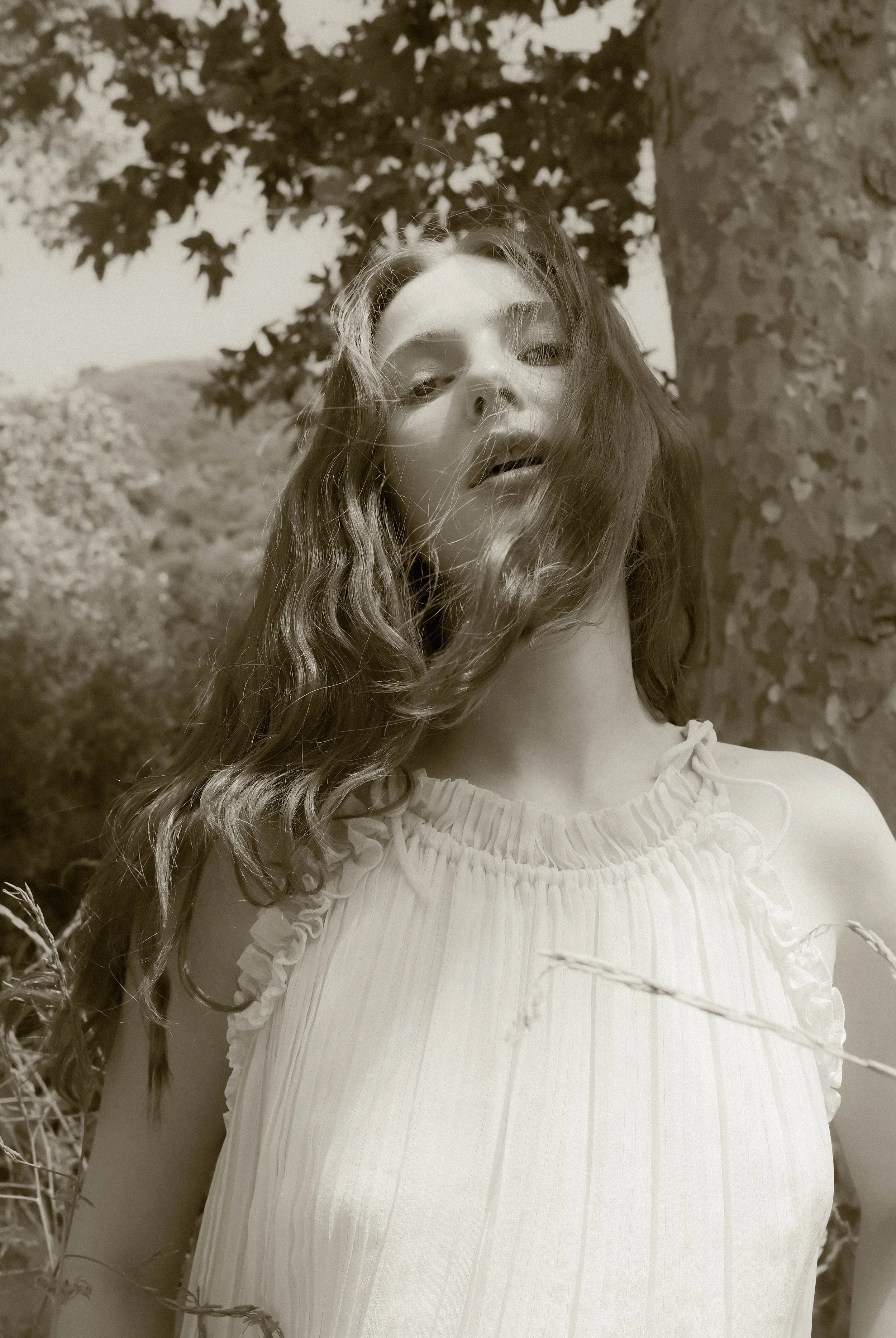 A woman with wavy hair looking at the camera, wearing a light-colored, ruffled sleeveless dress, outdoors with trees and foliage in the background, in black and white.