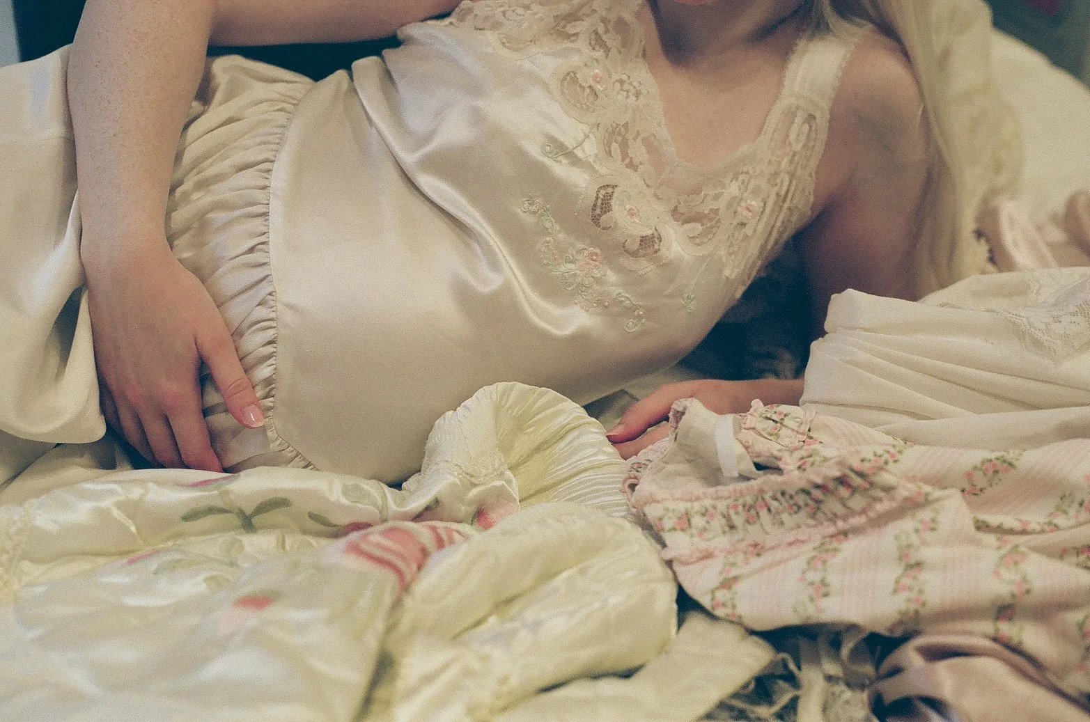 Close-up of a woman lying on bed, wearing a satin nightgown with lace details, surrounded by floral-patterned blankets and pillows.