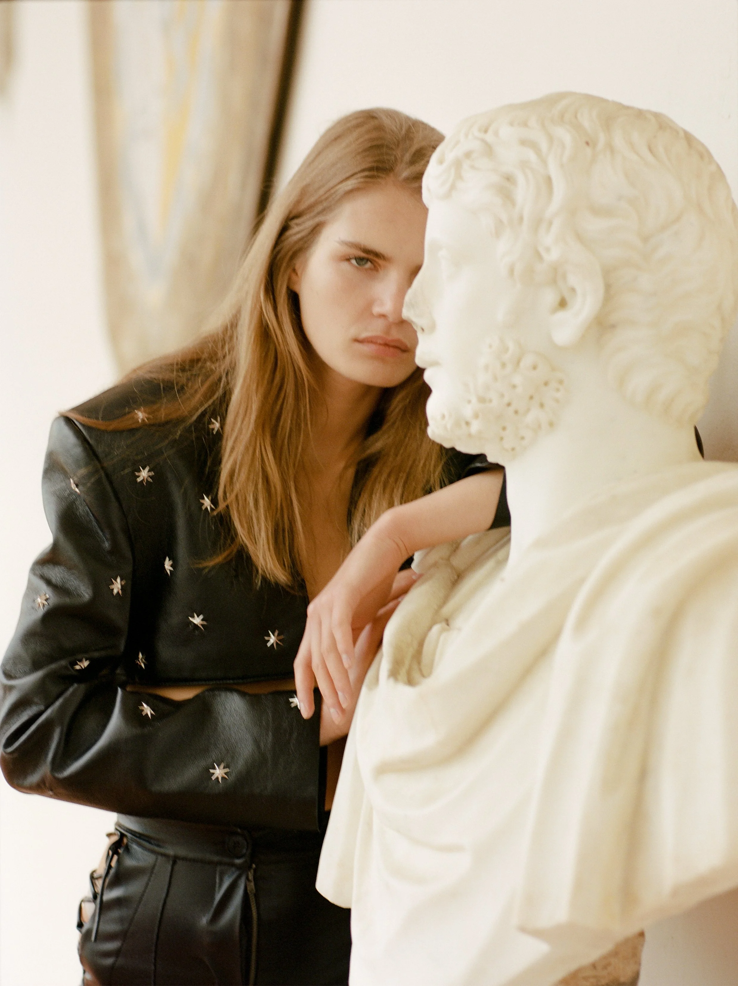 A woman with long, light brown hair poses closely next to a white marble bust of a male figure with intricate hair and beard details.