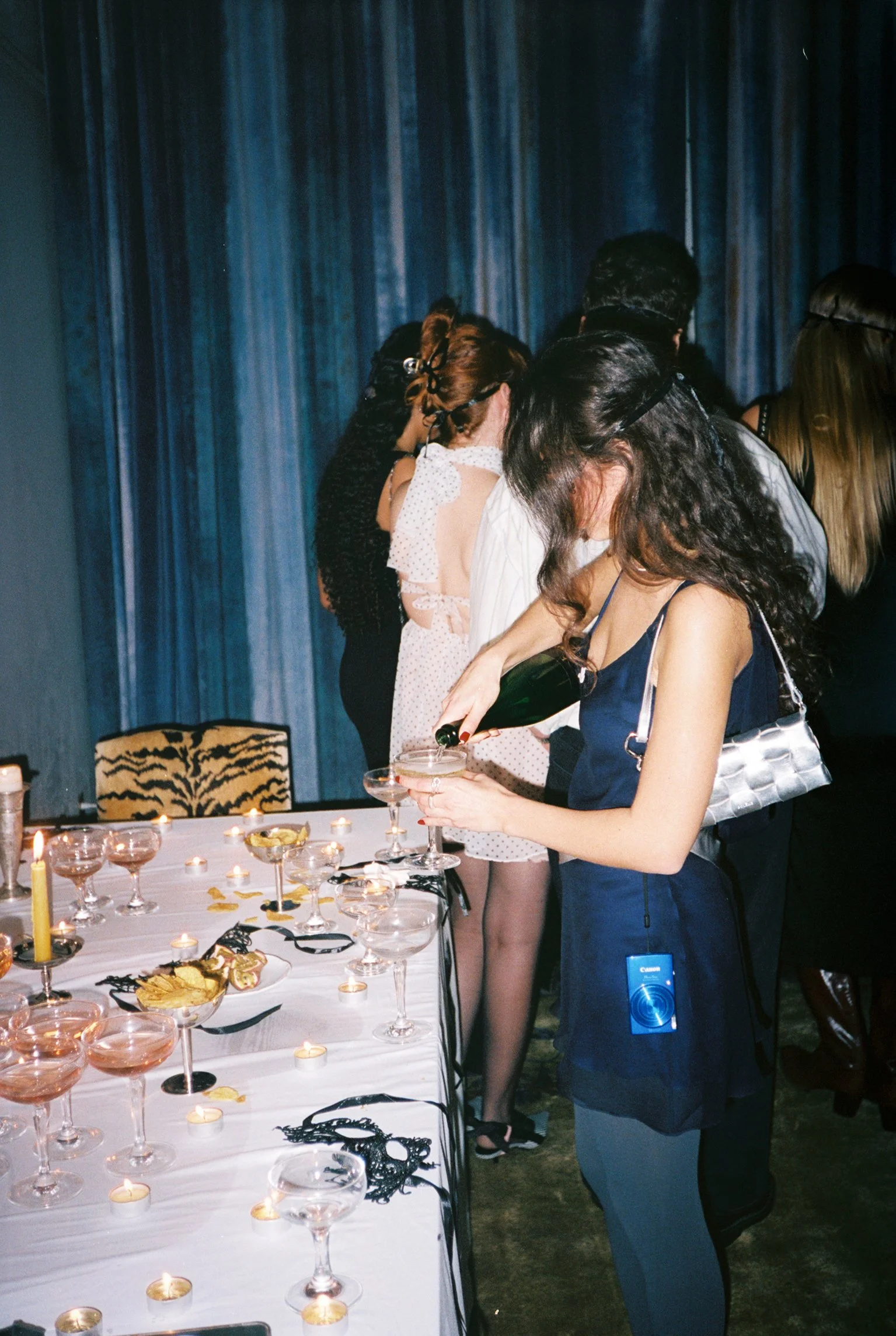 People standing around a table with candles and snacks, one woman pouring a drink from a bottle, dark blue curtains in the background