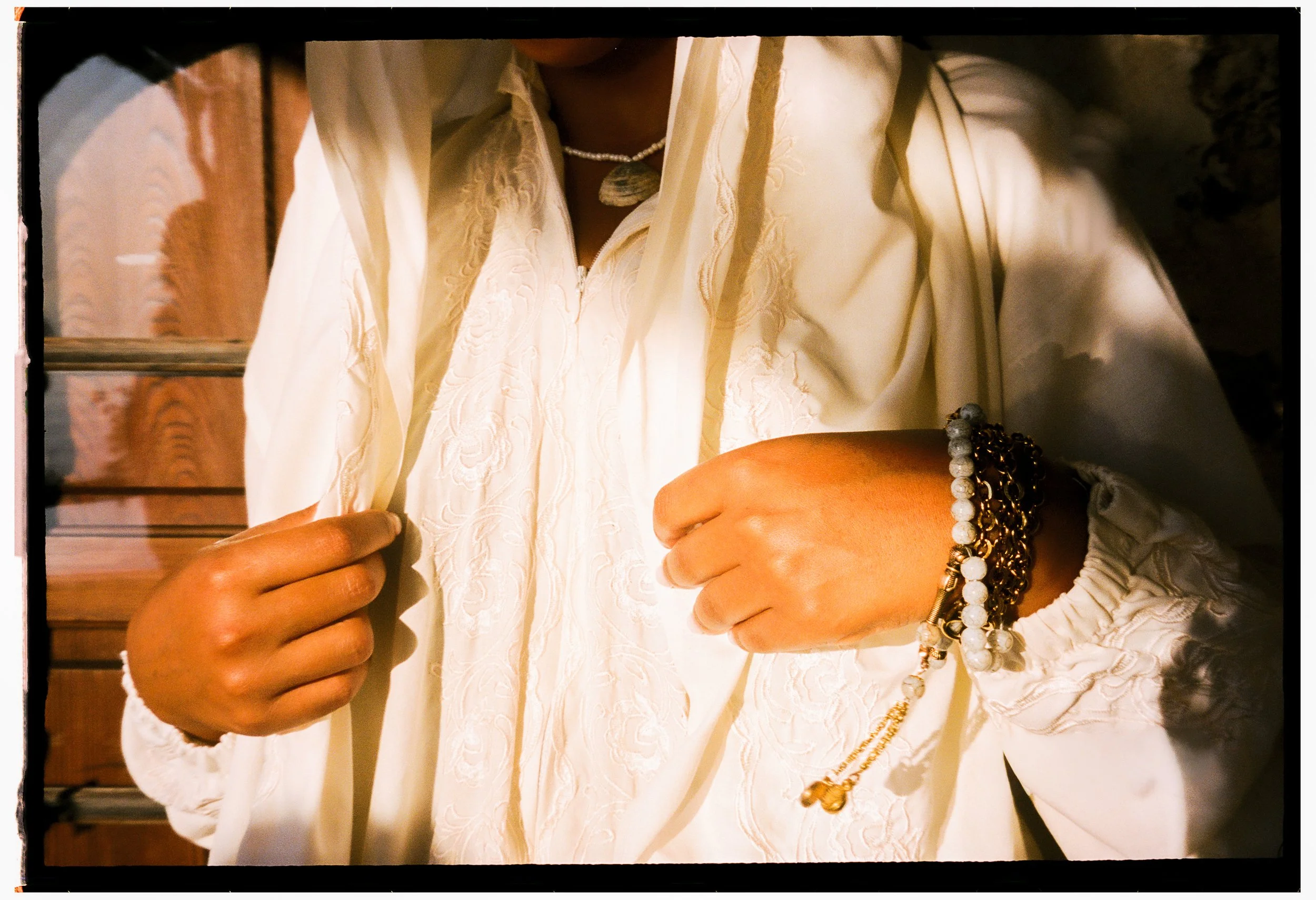 Close-up of a person adjusting a cream-colored embroidered garment, wearing layered jewelry including a pearl strand, a stone pendant necklace, and bracelets with beads and gold accents.