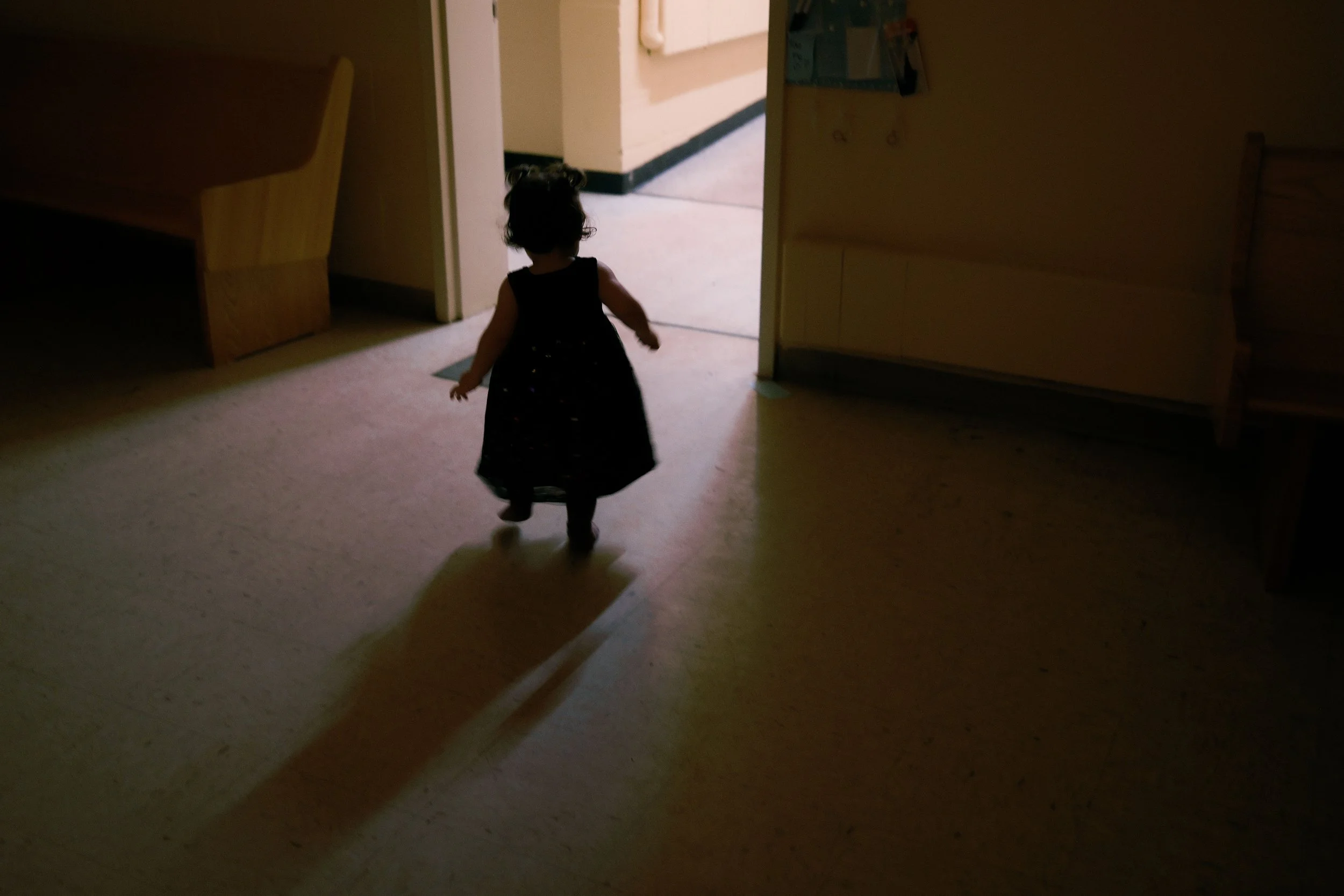 A young girl walking into a doorway in a dimly lit room, with a brightly lit area visible through the door.