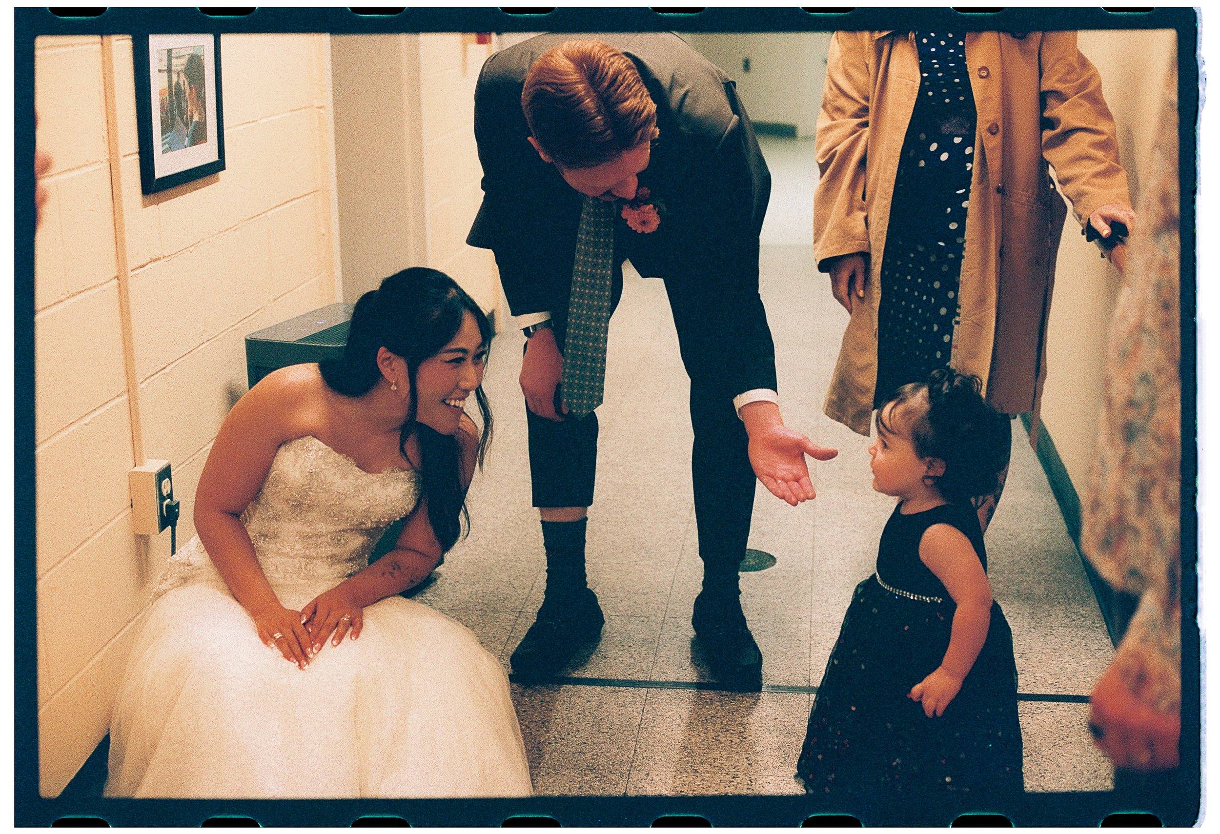 A bride smiling and sitting on the floor in her wedding dress while a man, dressed in a suit, is talking to a young girl in a black dress with polka dots. The setting appears to be in a hallway, with framed pictures on the wall and another woman stan
