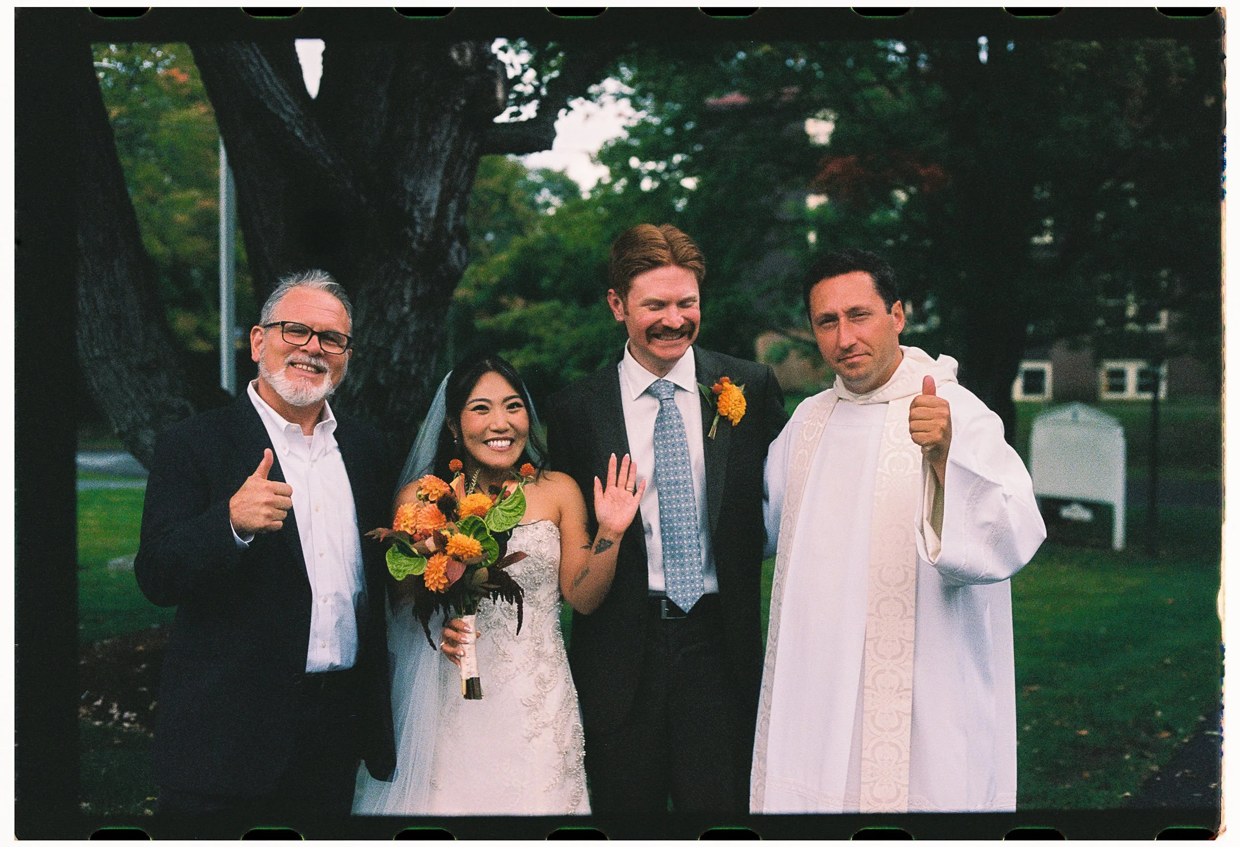 Group of five people standing outdoors in front of a large tree, including a bride holding a bouquet with yellow and orange flowers. The group appears happy, with some smiling and one giving a thumbs-up, possibly at a wedding or celebration.