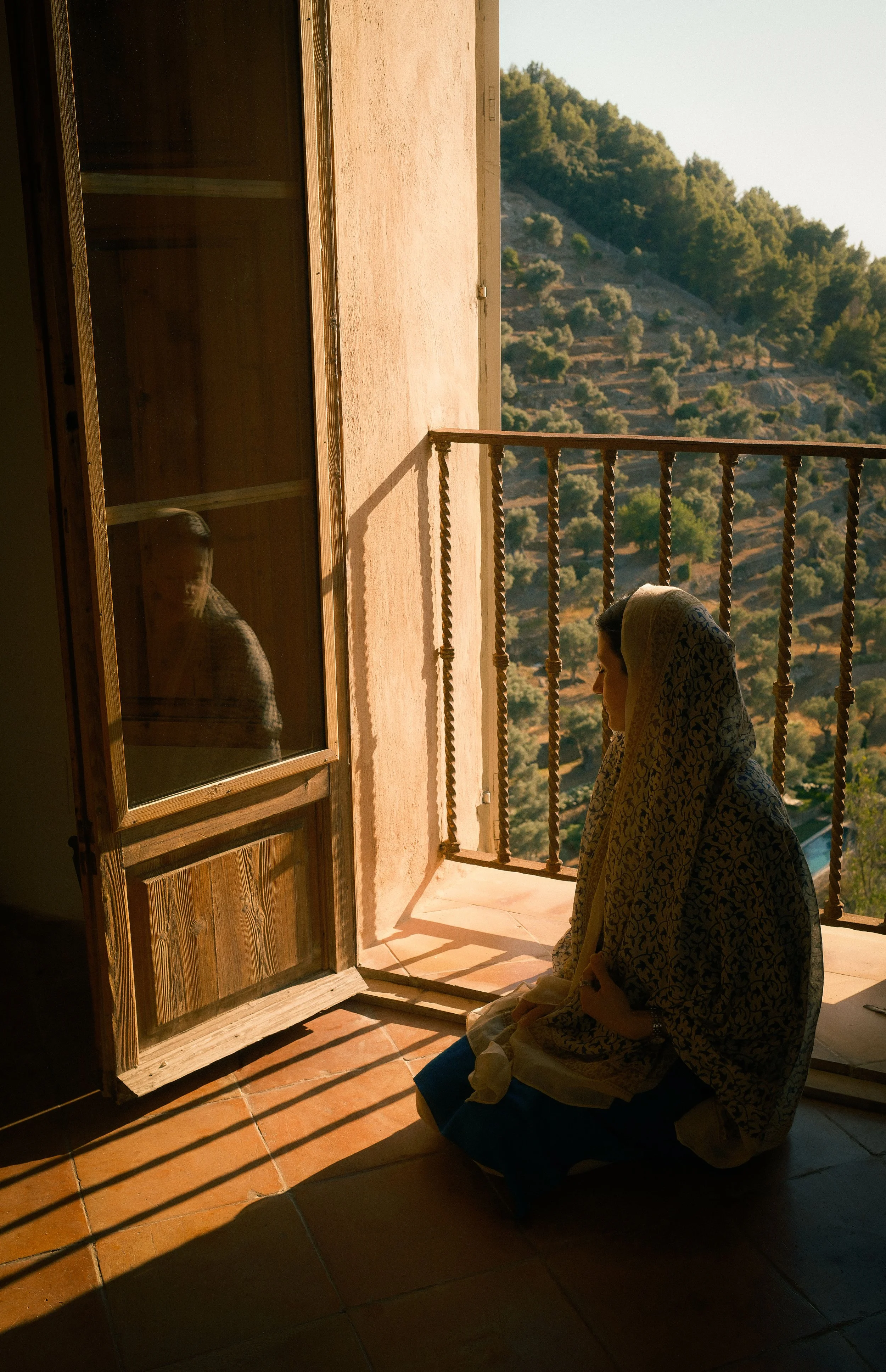 Two women, dressed modestly with headscarves, are sitting on the floor on a tiled porch or balcony, facing a landscape of hills covered in trees. One woman is sitting with her eyes closed. They are illuminated by warm sunlight, casting shadows on the