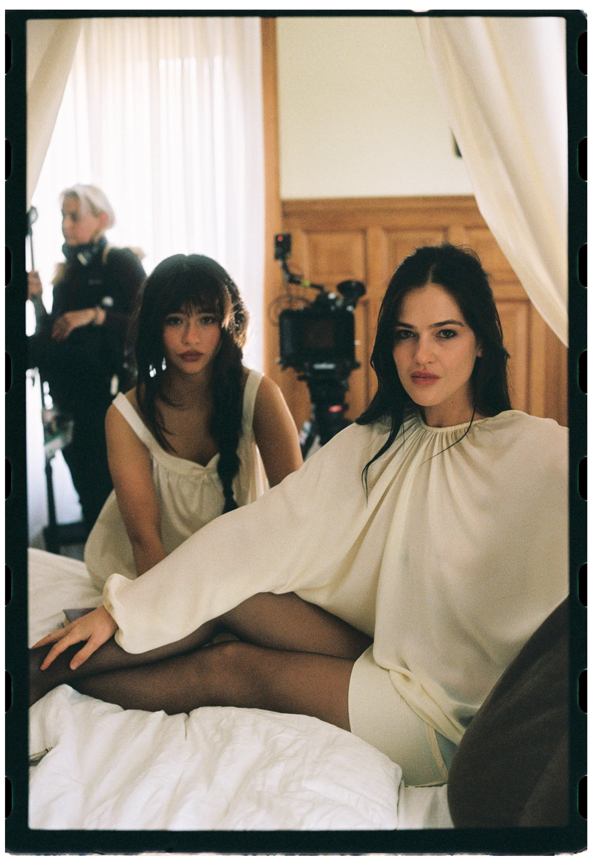 Two women sitting on a bed, one with dark hair and a white dress, the other with dark tattoos and a light top, with woman in background adjusting her hair, in a hotel room with wooden paneling and curtains.