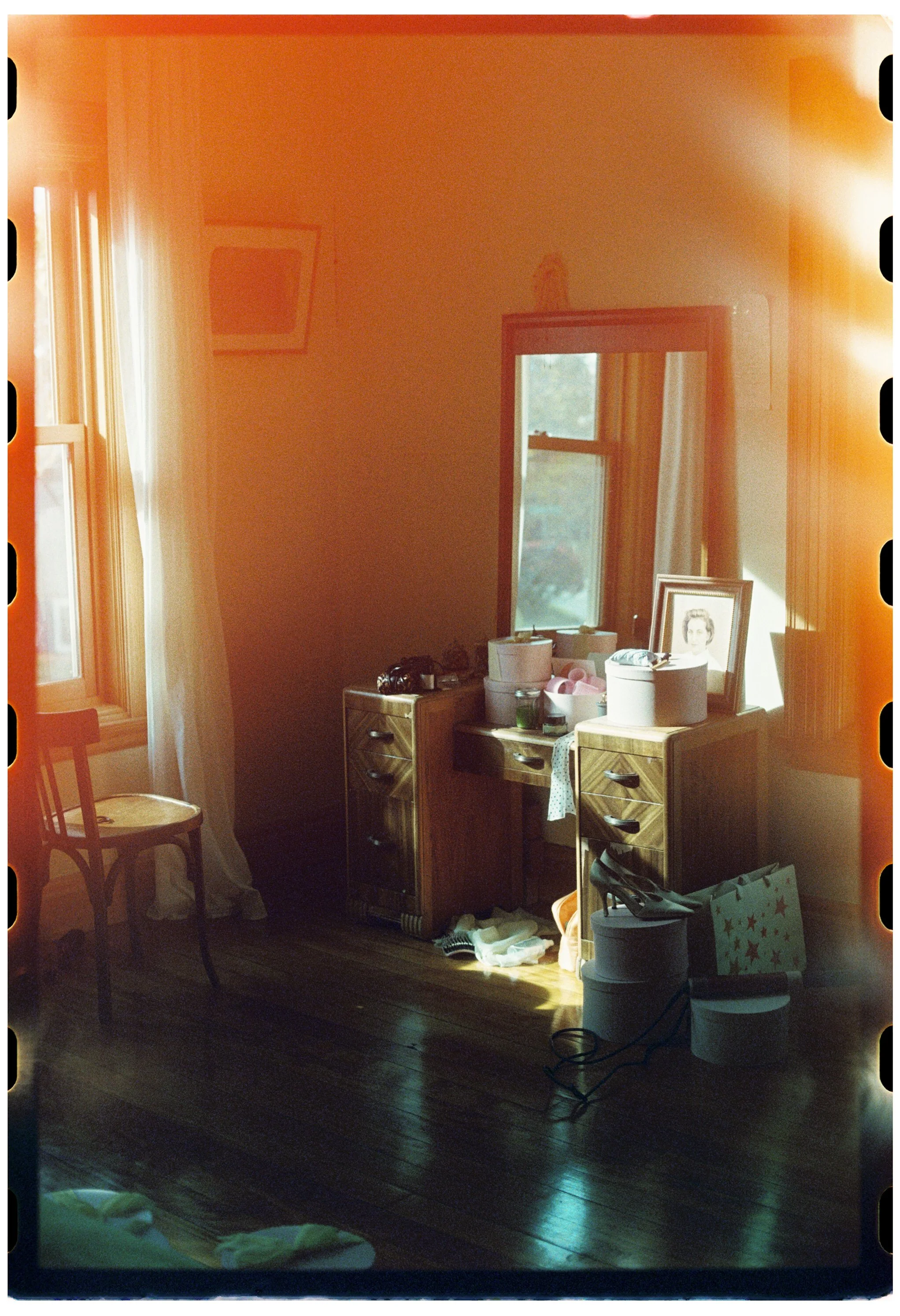 A cozy bedroom corner with wooden furniture, a window with curtains, a framed photo on a dresser, and various household items, bathed in warm sunlight.