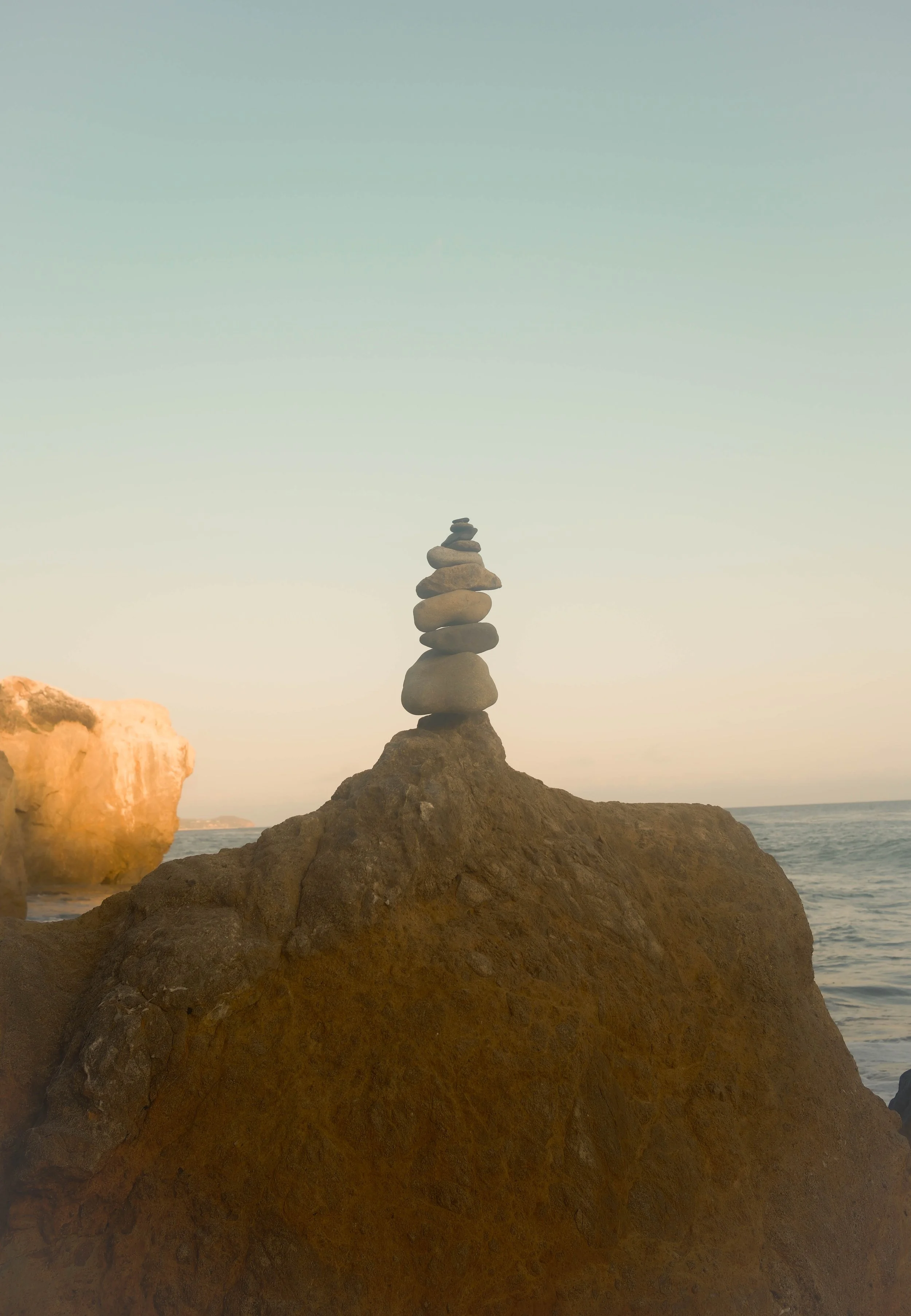 Stacked rocks on a large rock near the beach with ocean waves in the background during sunset.