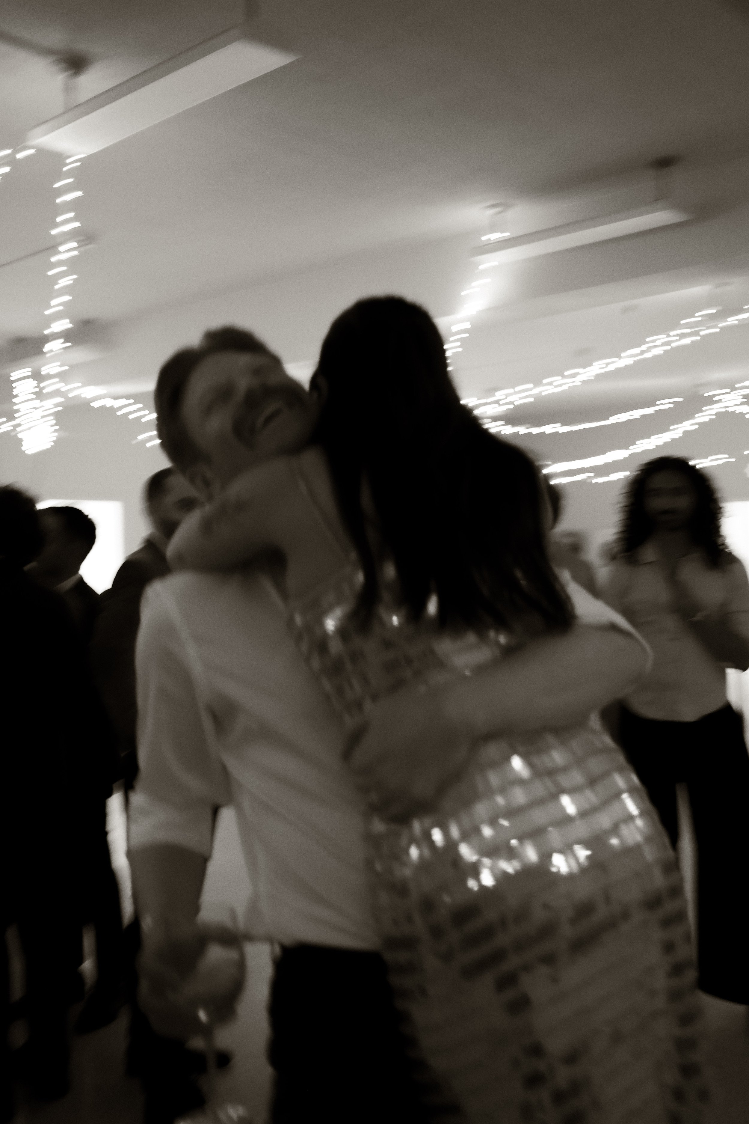 People gathered at a celebration or party, with festive lights hanging from the ceiling, some individuals hugging and smiling.