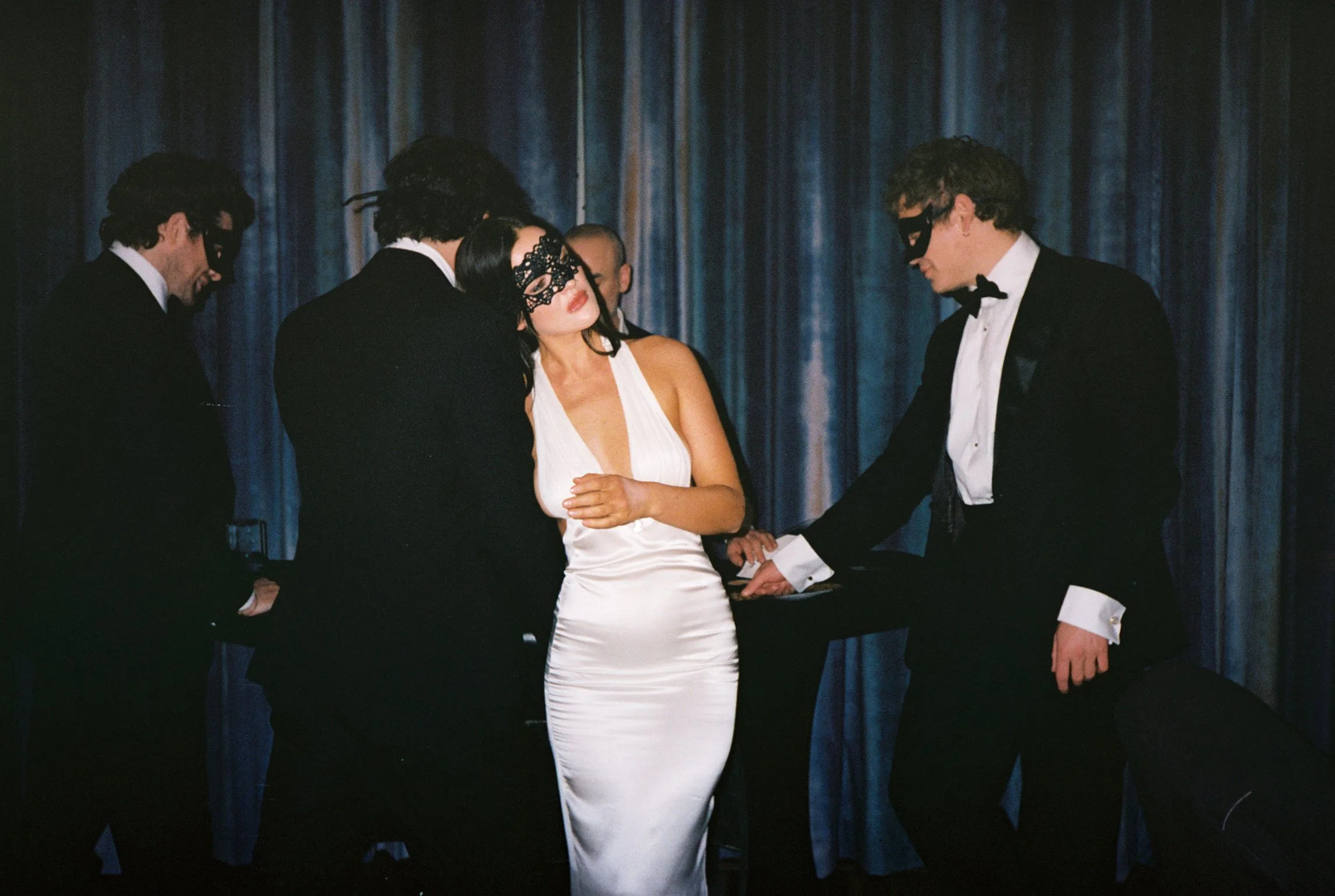 A group of five people dressed in formal attire, wearing black masks, engaged in a ceremony or ritual, on a stage with dark blue curtains in the background.