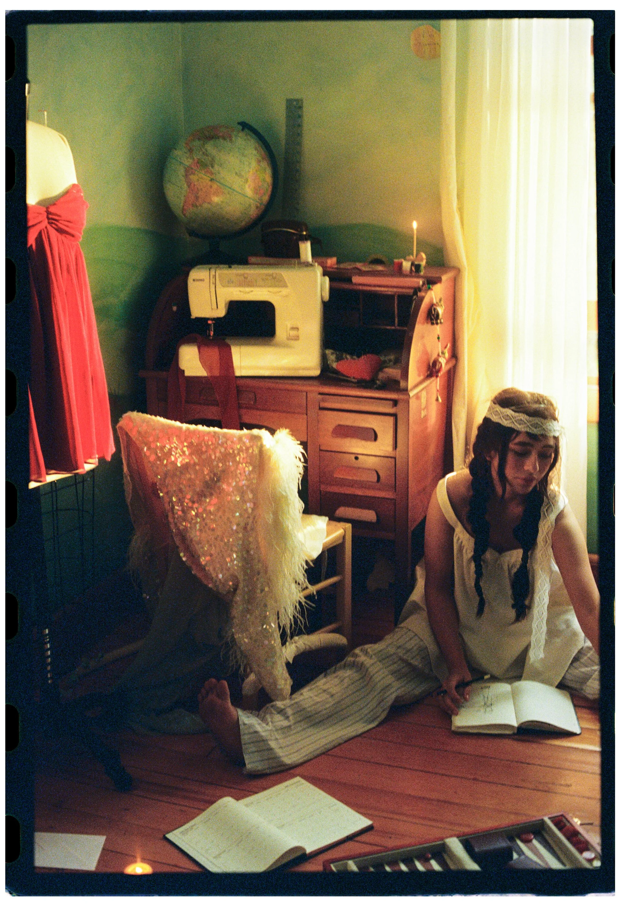 A young woman with long braided hair, wearing a white dress with lace details, sits on the floor reading a book in a cozy room with natural light. Behind her, there is a desk with a globe, a lamp, and a sewing machine. A glittery high-heeled shoe wit