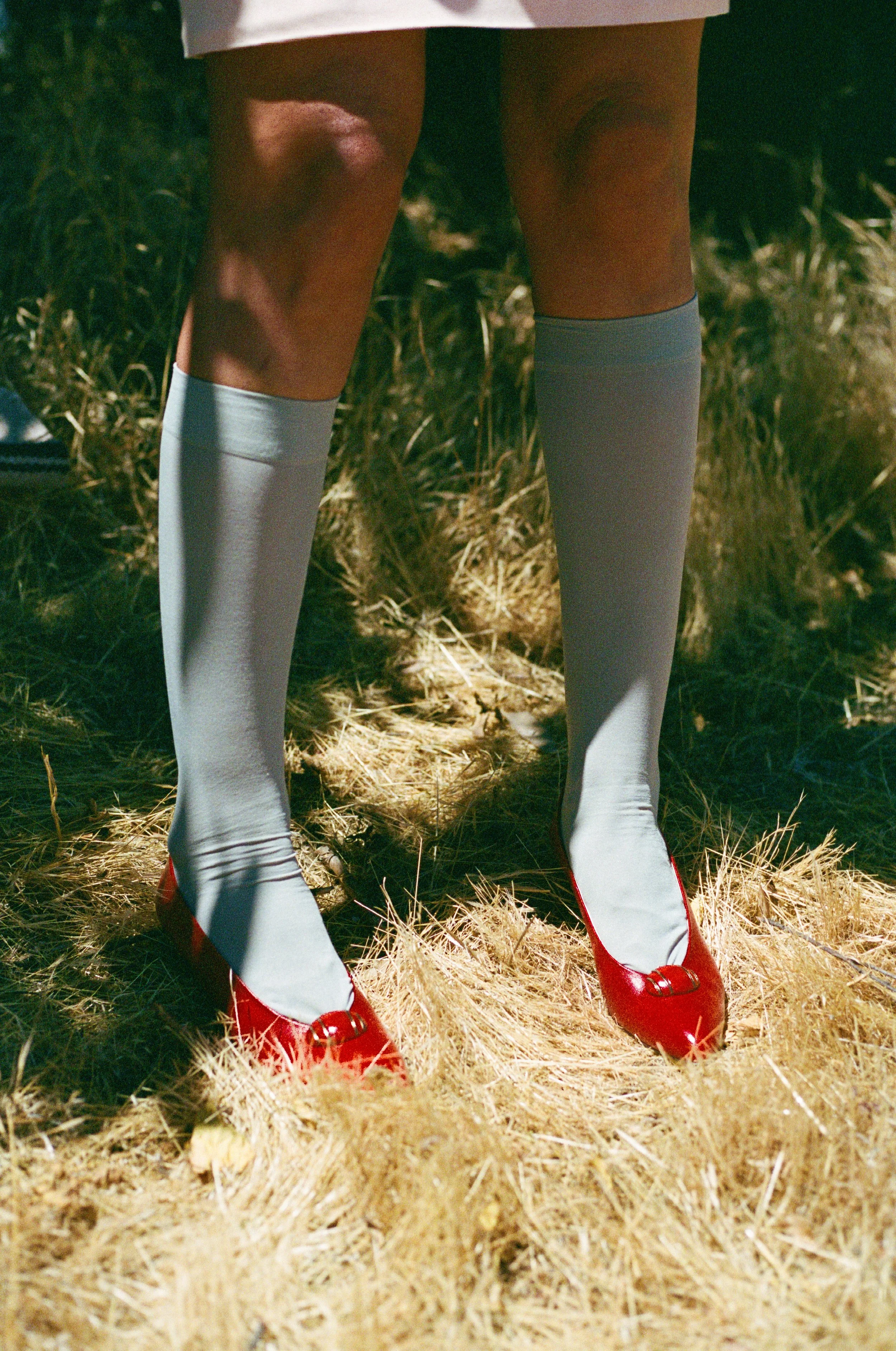 Close-up view of a person wearing white thigh-high socks with red shoes and a white skirt, standing on dry grass outdoors with greenery in the background.
