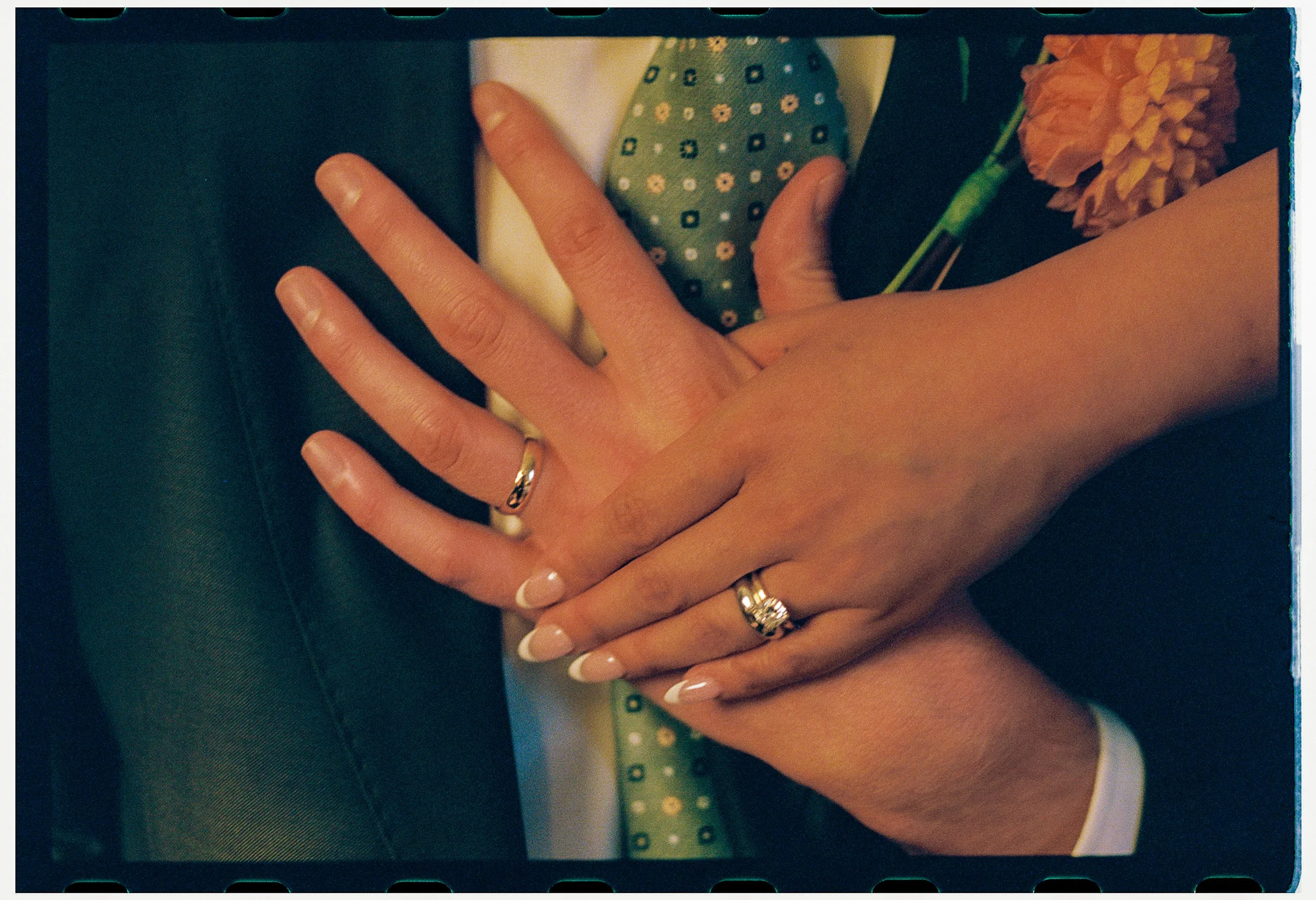 Close-up of two hands with rings, one hand resting on top of the other, on a surface with a green patterned object and a pink flower in the background.