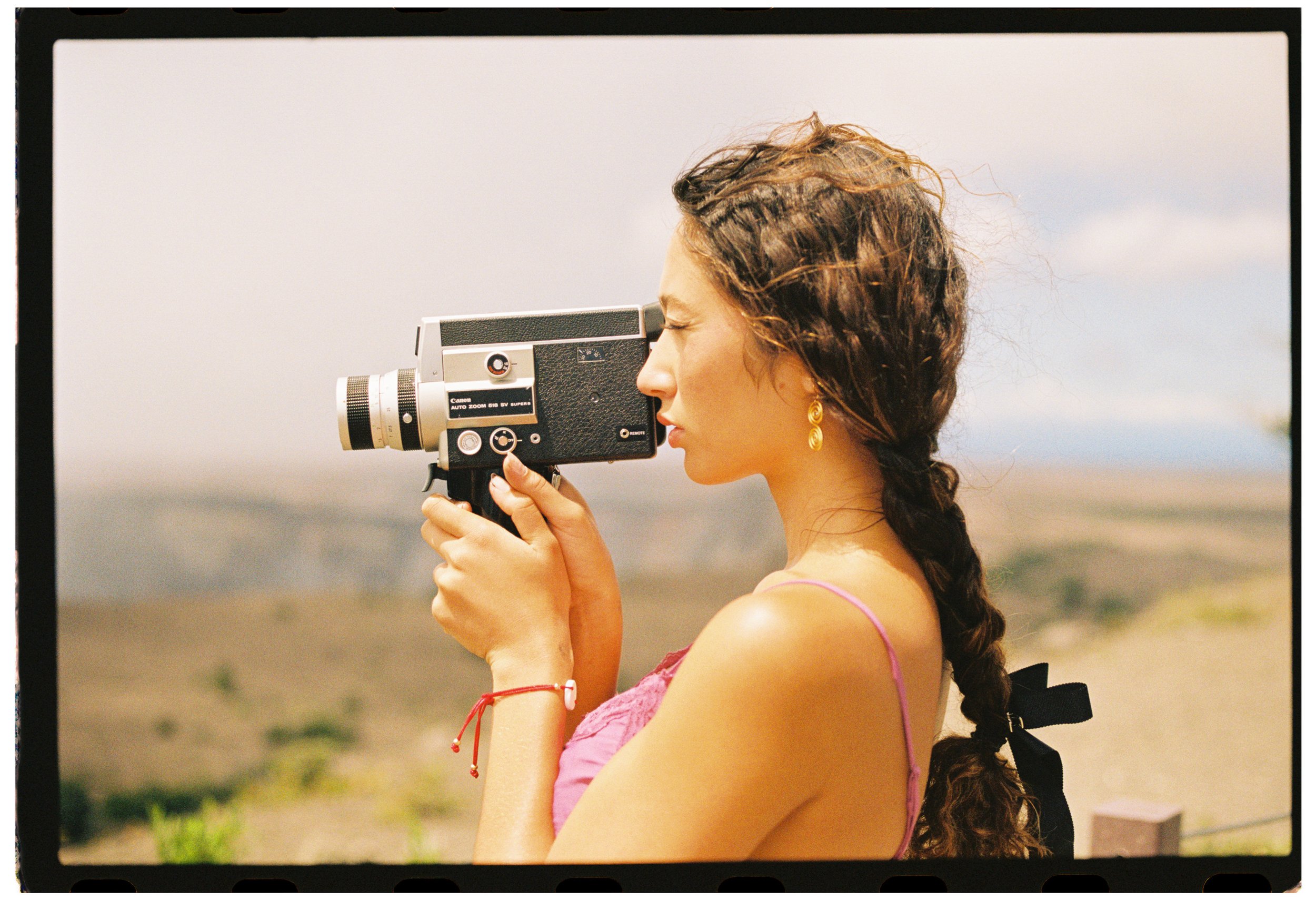 Woman with curly hair and earrings taking a photograph outdoors using a vintage camera, with a blurred natural background.