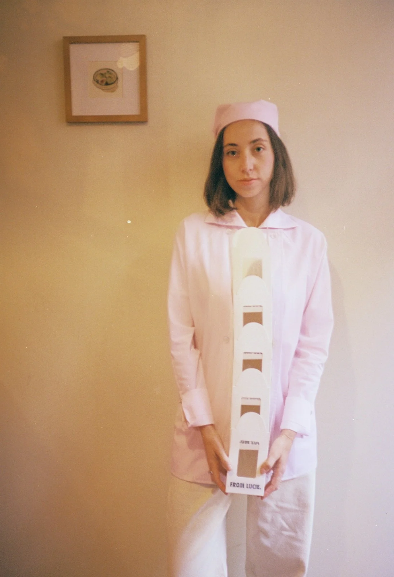 A woman in pink pajamas and a matching pink sleep cap is standing against a beige wall, holding a tall white object with black stripes labeled "FROM LUCCE." There is a framed picture of food on the wall behind her.