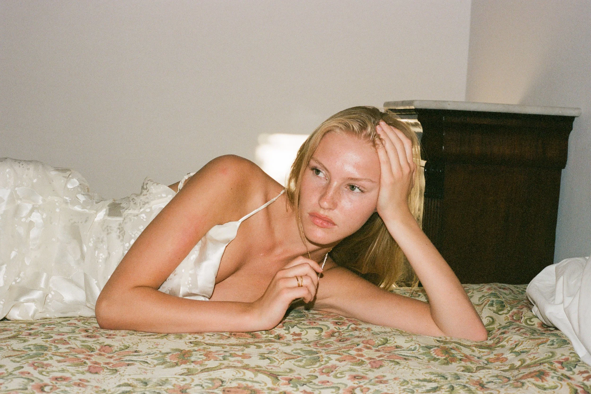A young woman with long blonde hair lying on a floral patterned bed, resting her head on her hand, wearing a white spaghetti strap dress, with a pensive expression, in a bedroom setting.