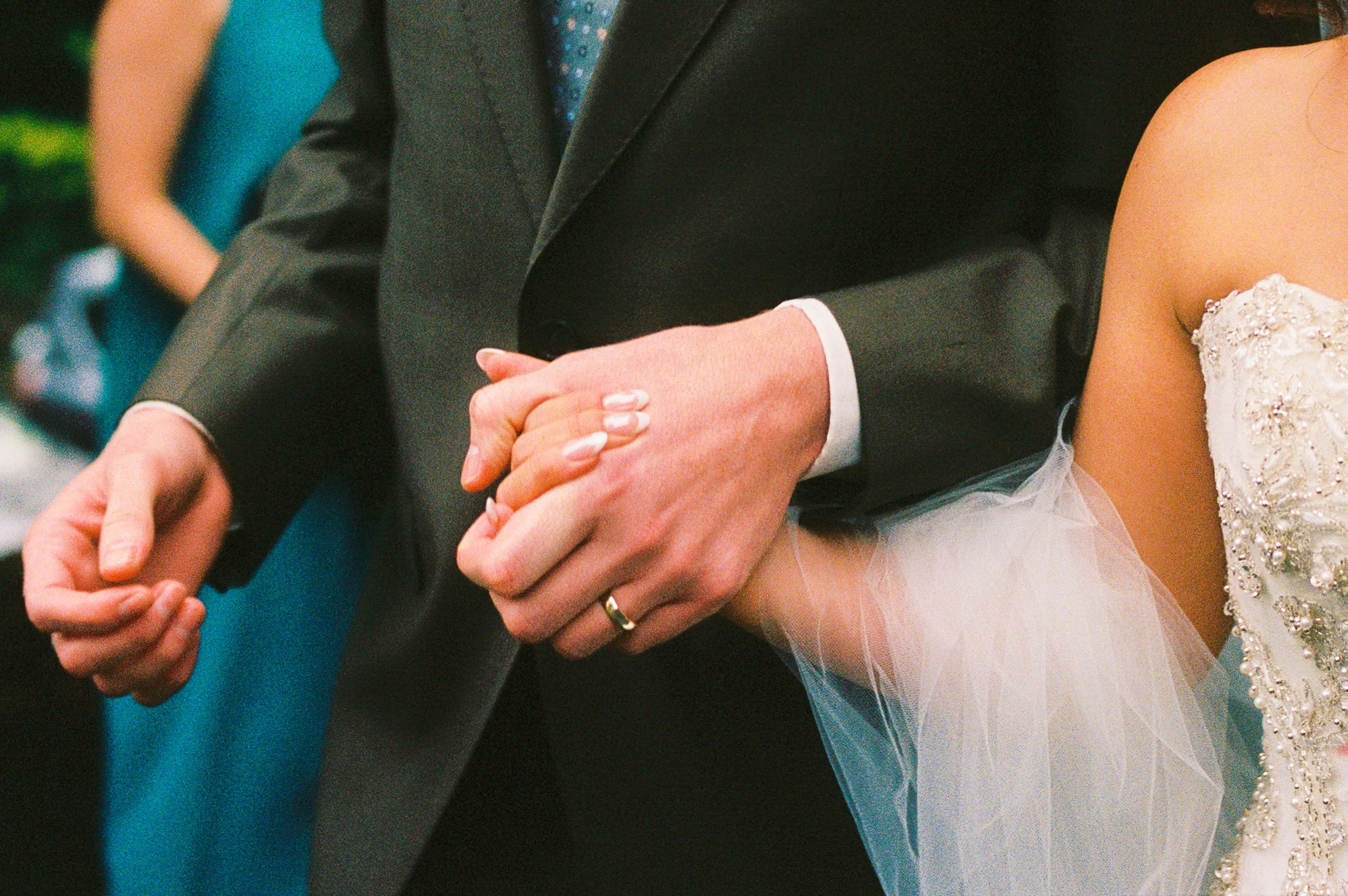 Close-up of a couple holding hands during a wedding ceremony, with the bride wearing a white dress and the groom in a black suit.