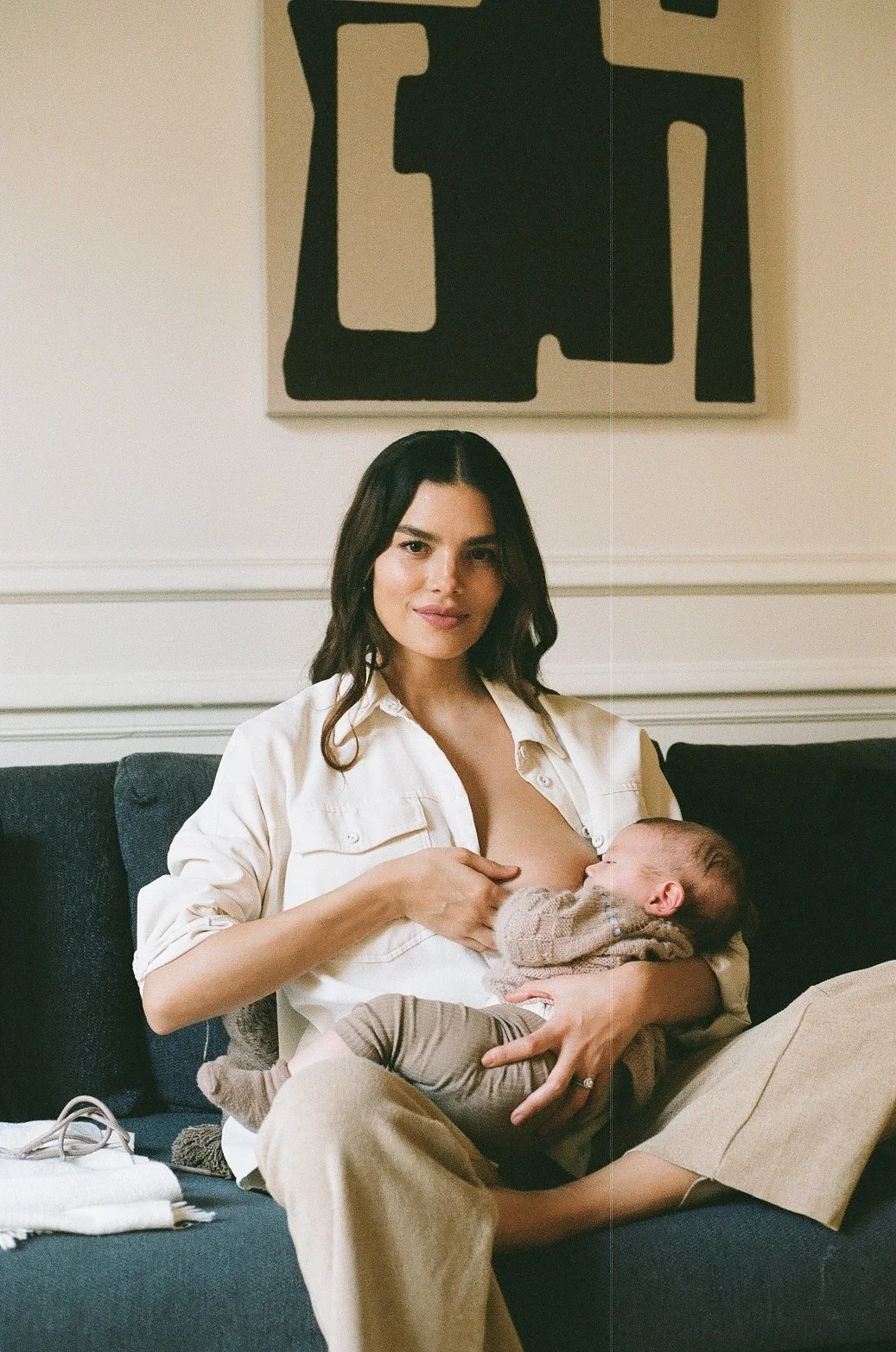 A woman in a white shirt is breastfeeding a baby with a knit sweater, sitting on a dark couch with a pillow, in a room with cream walls and abstract artwork hanging behind her.