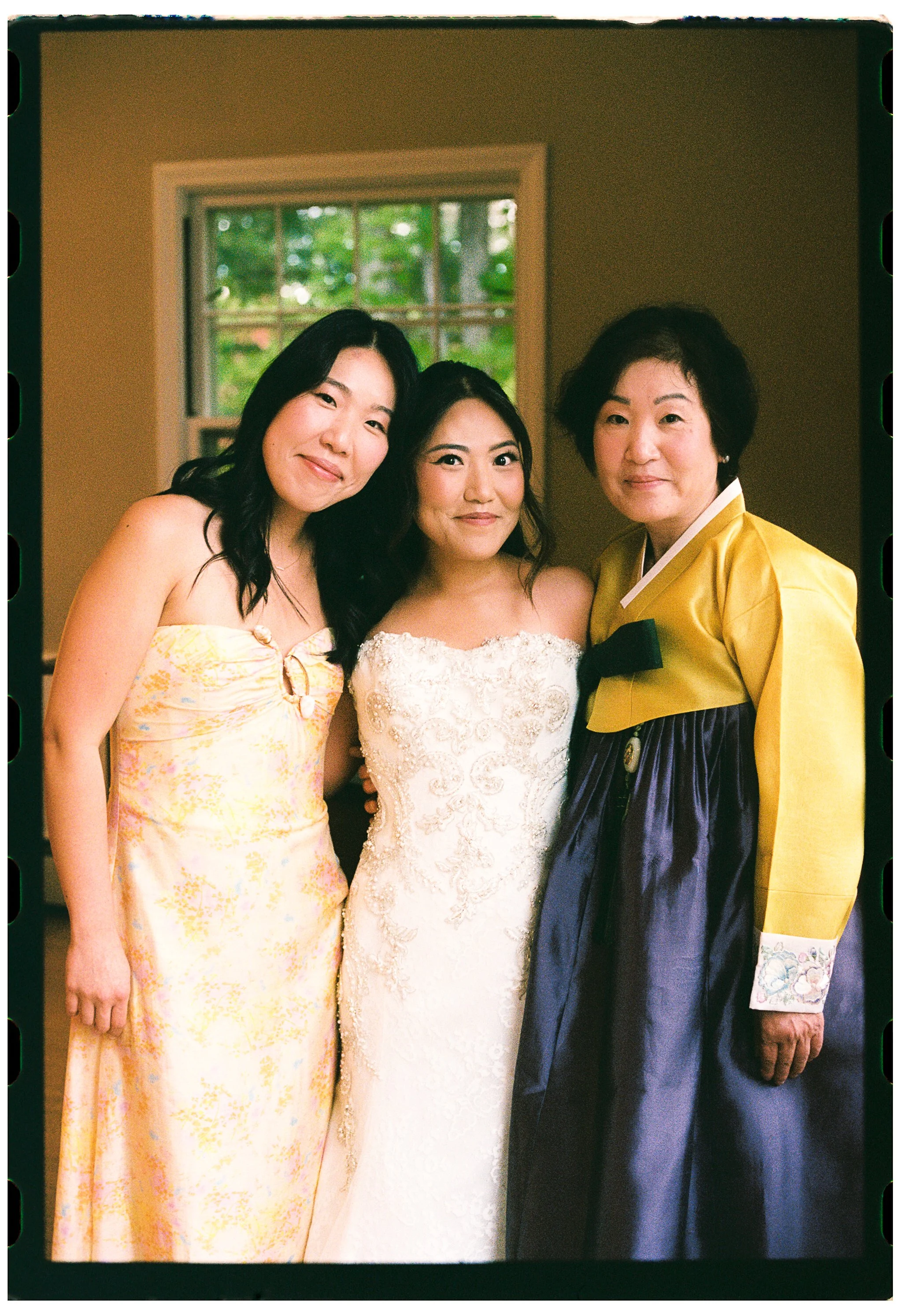 Three women standing close together indoors near a window with greenery outside. One woman is wearing a yellow and white dress, the woman in the middle is in a white lace dress, and the third woman is in a yellow jacket with a black ribbon. All three