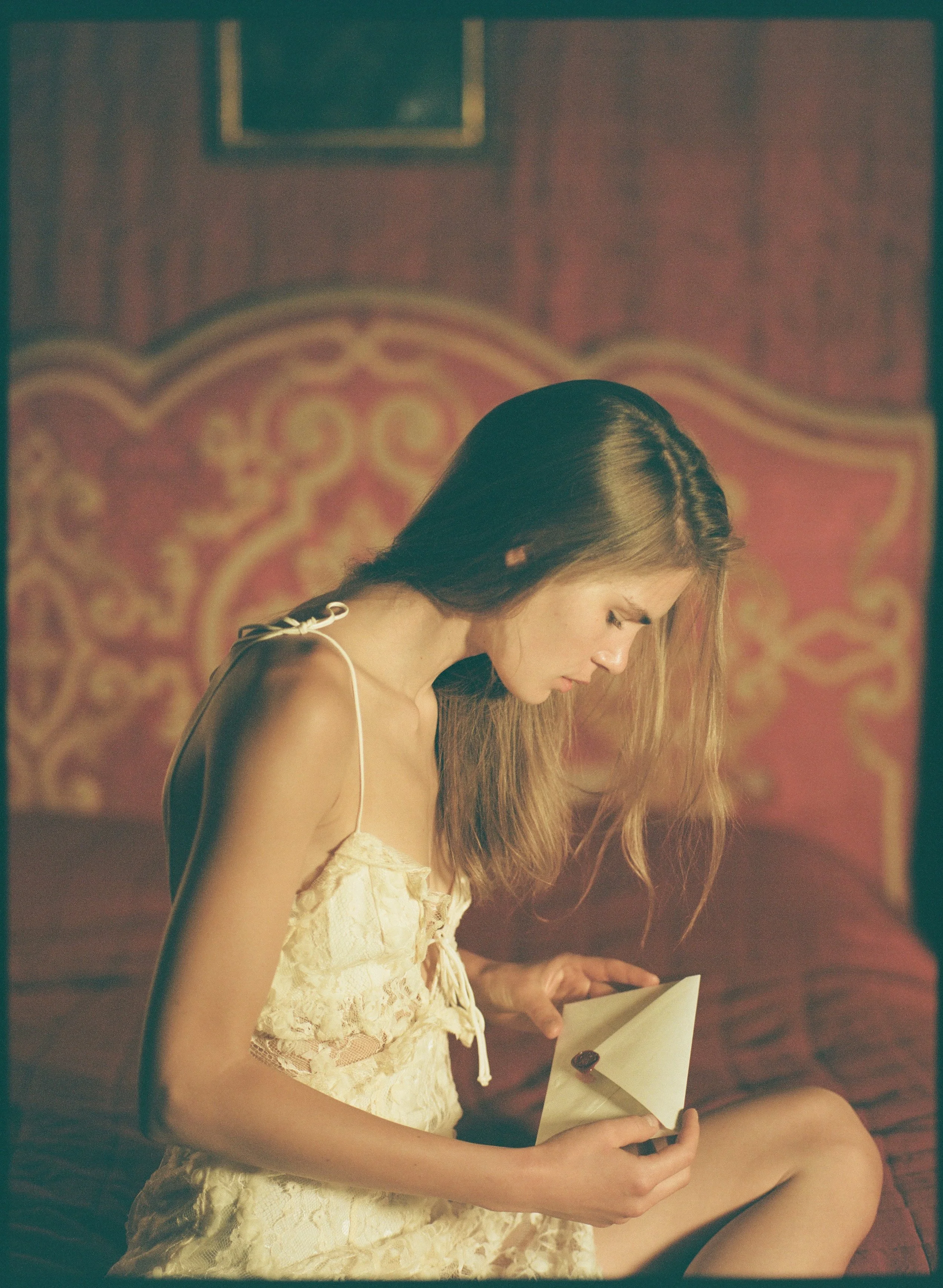A young girl with long hair wearing a yellow lace dress, reading a letter while sitting on a bed with an ornate headboard and framed pictures in the background.
