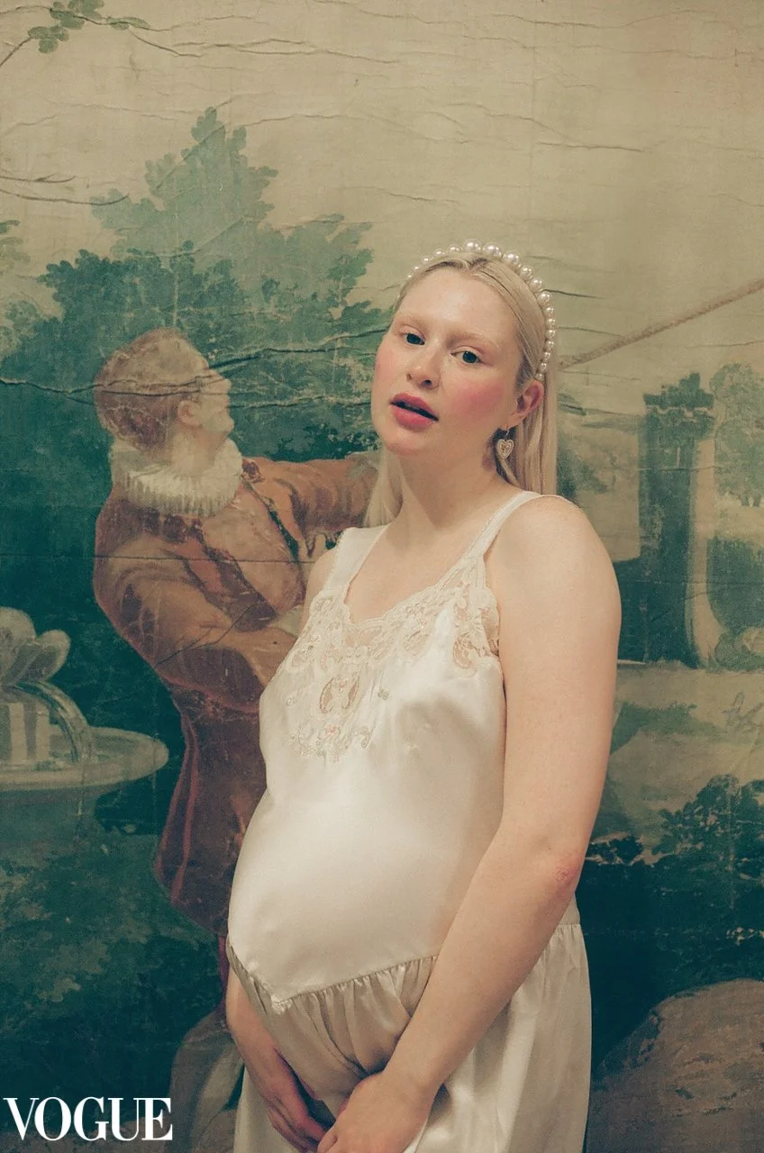 A young woman with light blond hair wearing a pearl headband and cream-colored satin dress with lace details, standing in front of a painted mural of a historical scene with a woman in period dress and a landscape in the background.