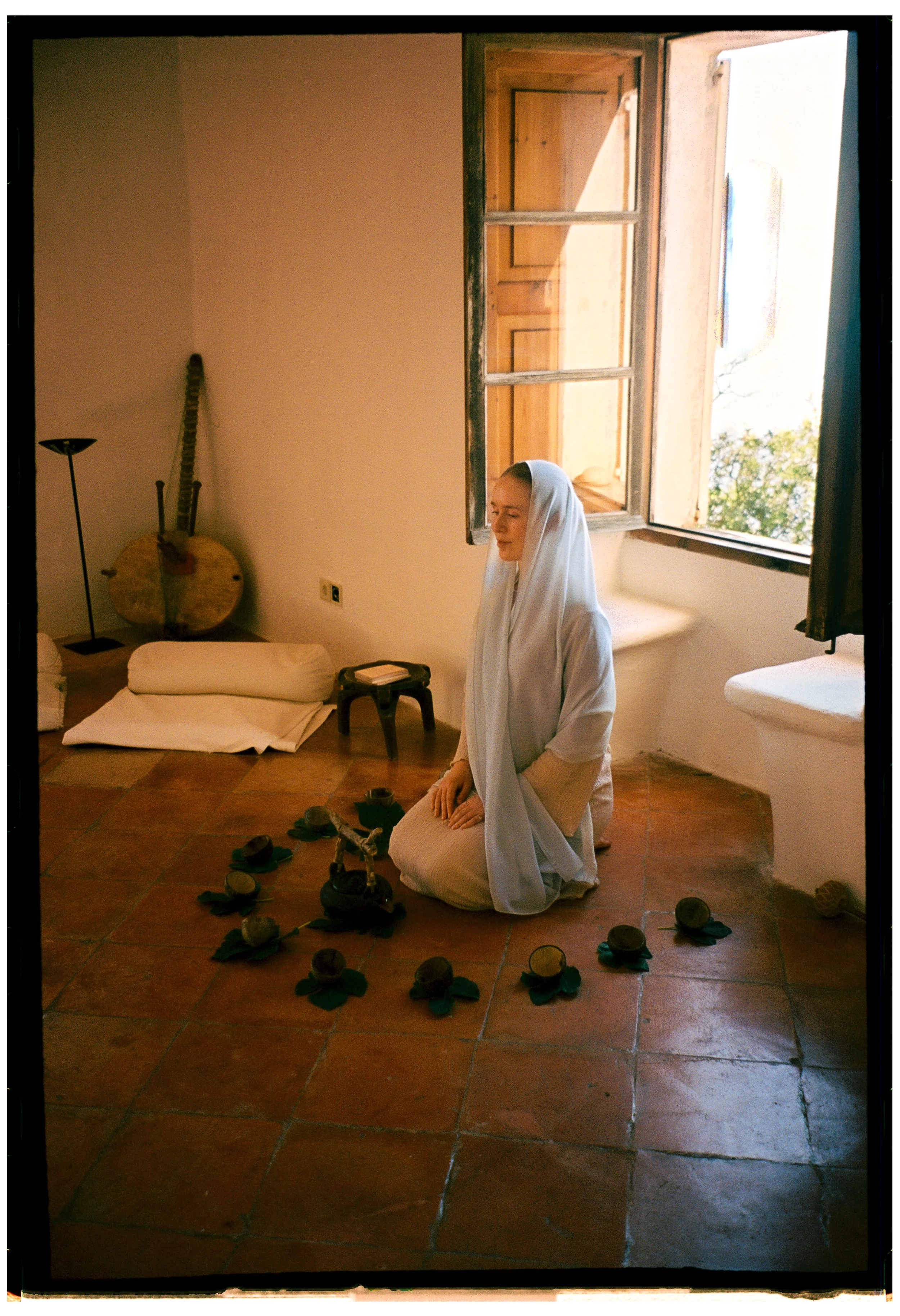 A woman kneeling on the floor with her eyes closed, wearing a white head covering and robes, surrounded by scattered walnuts with green leaves, in a room with open windows letting in natural light, and various objects including a folded blanket, a bl