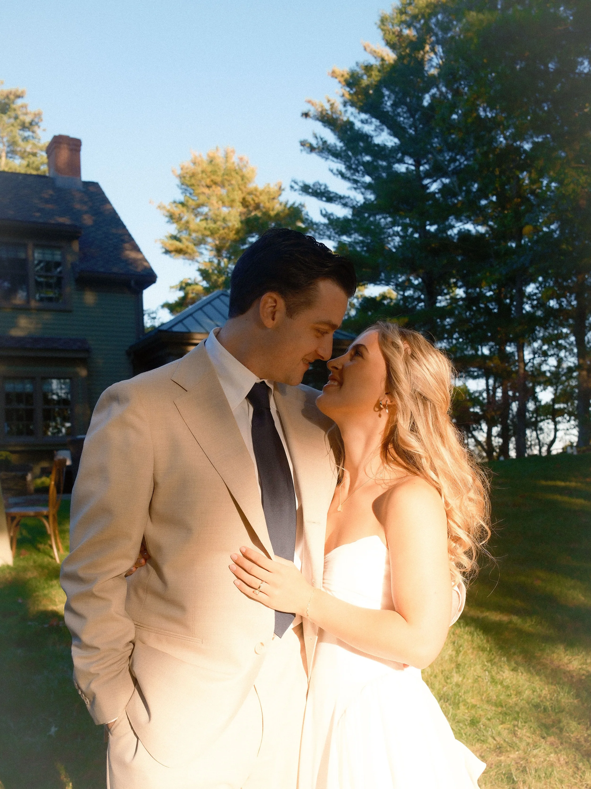A newlywed couple share an intimate moment outdoors during sunset, with trees and a house visible in the background. Shot on 35mm film. Cape cod wedding golden hour