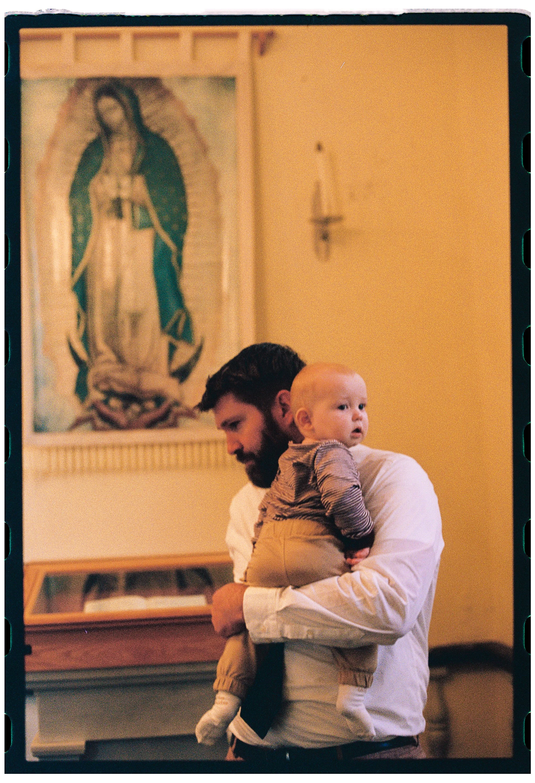 A man with dark hair and a beard holding a young child with blonde hair inside a room with religious artwork on the wall, including a large image of Our Lady of Guadalupe.