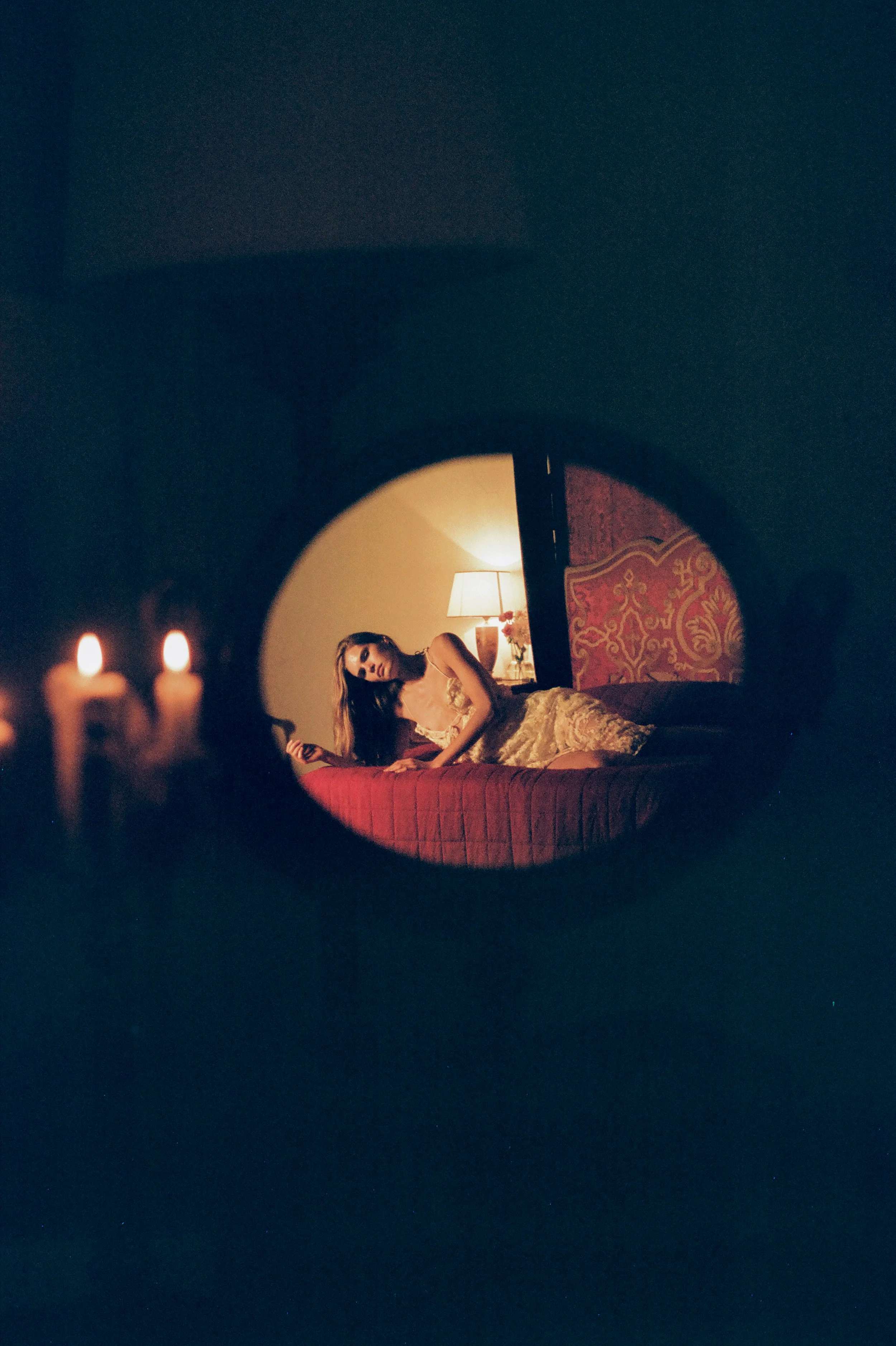 A woman lying on a bed in a dimly lit room, with a lamp on a nightstand behind her, visible through a mirror reflection. Two candles are in the foreground.