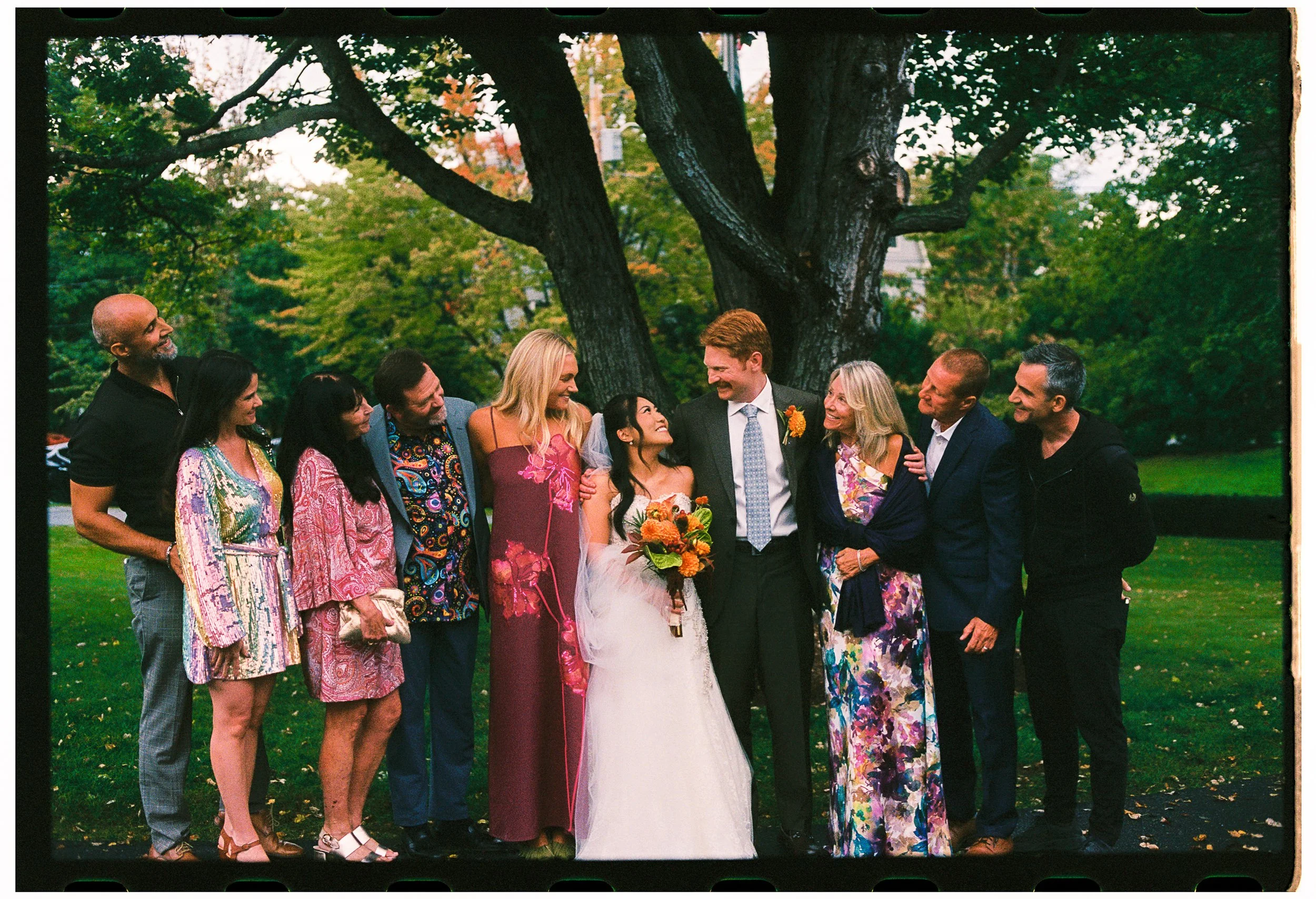 Group of people gathered outdoors around a bride and groom, smiling and looking at each other, with trees and green grass in the background.