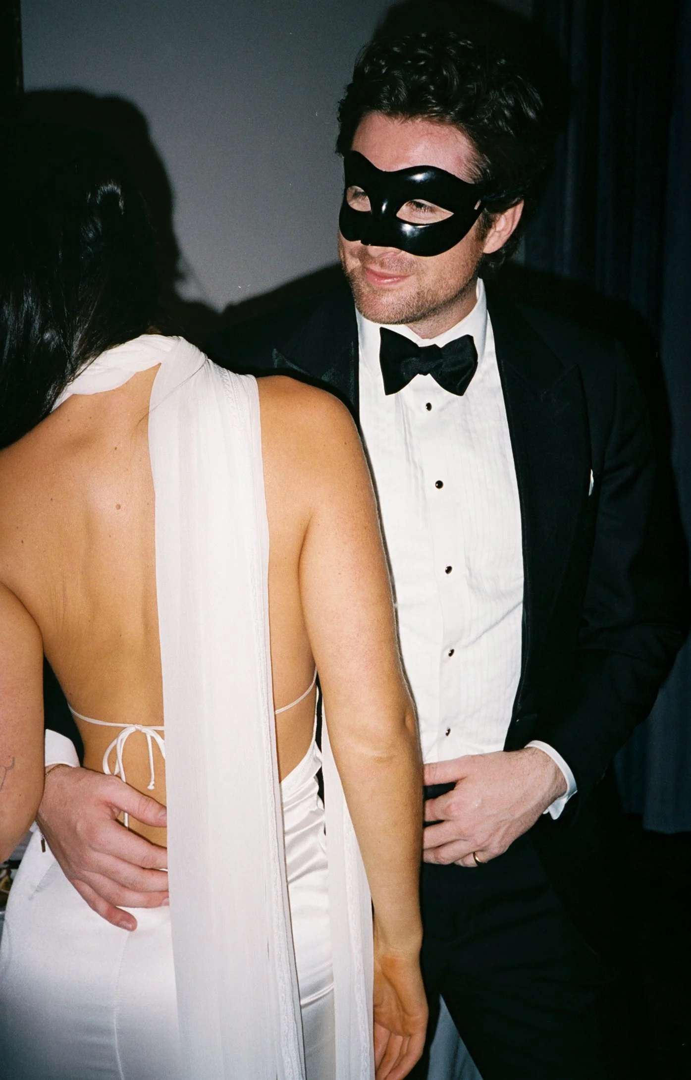 Close-up of a man in a tuxedo and black mask, holding a woman in a white dress at a formal event.