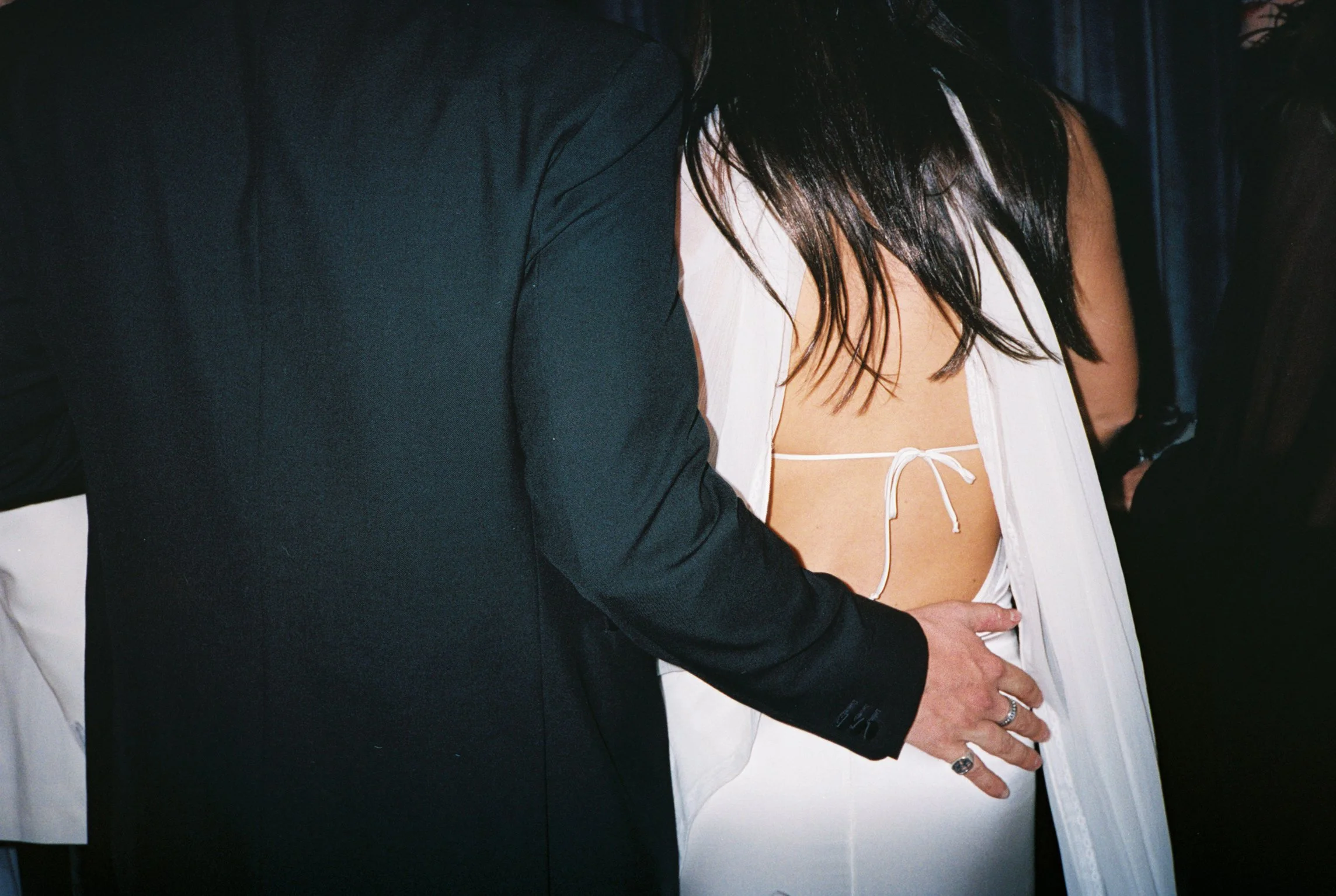 Close-up of a couple embracing, with the focus on the woman's back and the man's arm around her waist. The woman has long dark hair and is wearing a white dress, while the man is dressed in a dark suit.