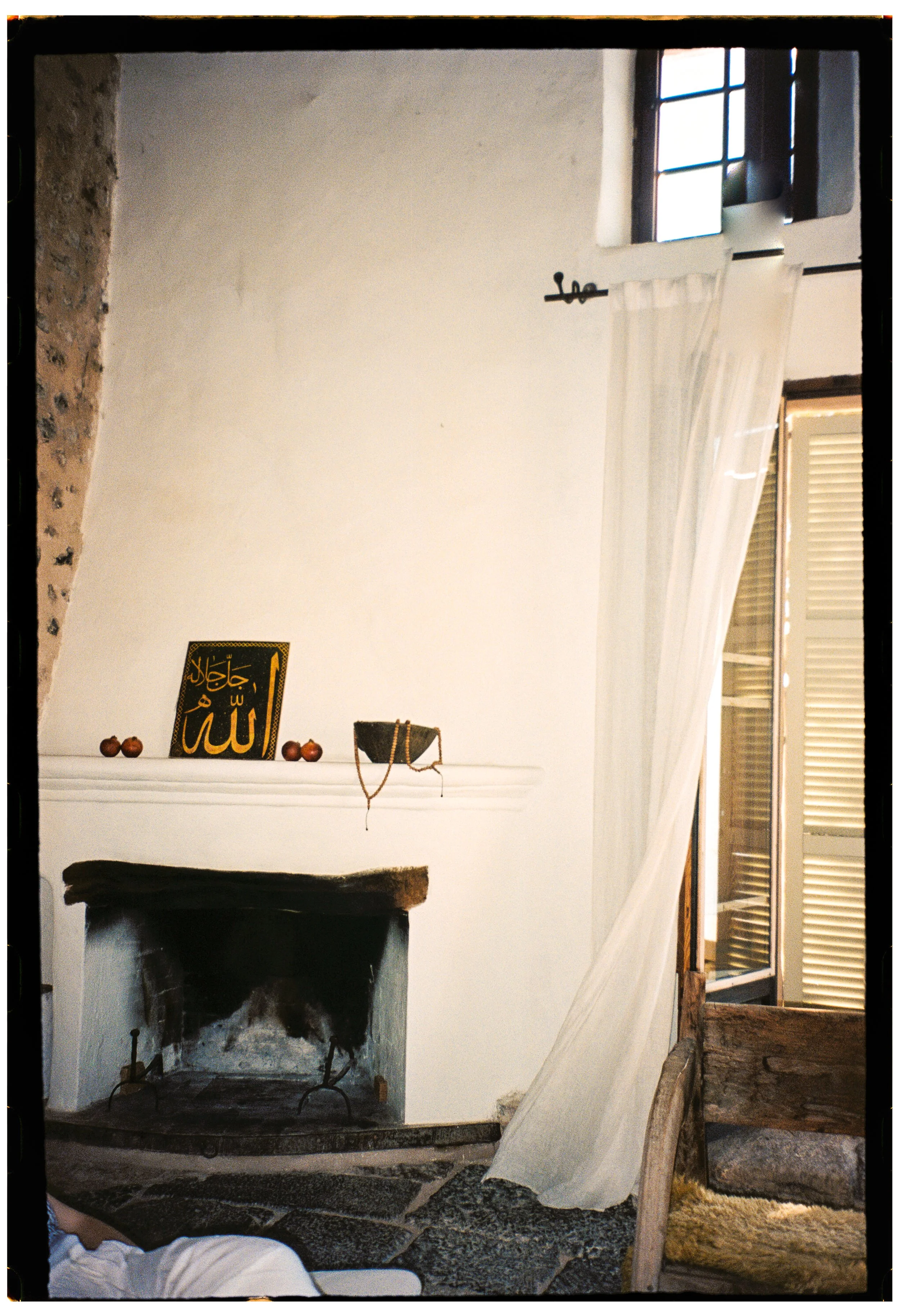 Interior view of a room with a fireplace, a small shelf with decorative objects, a framed Islamic calligraphy piece, and a window with open shutters and sheer curtains.
