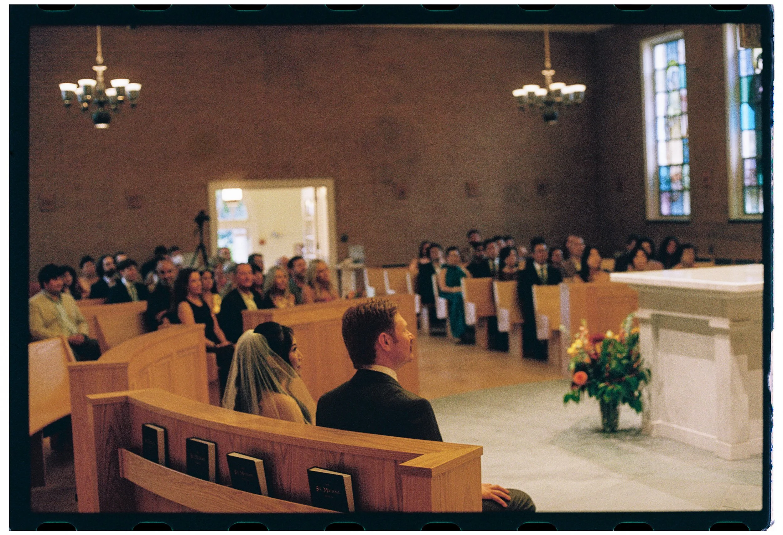 People seated in wooden pews inside a church during a ceremony, with stained glass windows and chandeliers hanging from the ceiling.
