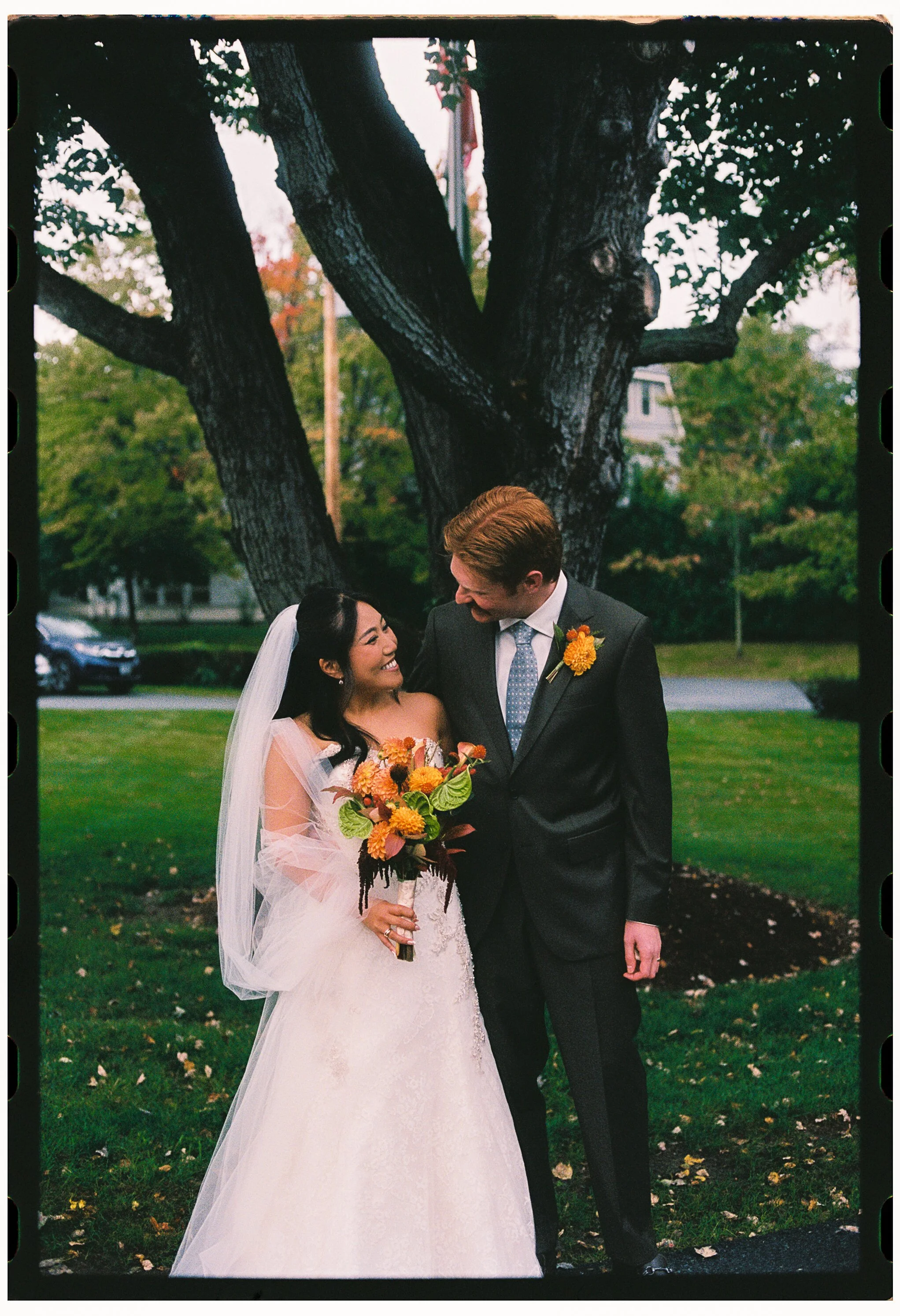 A newlywed couple standing outdoors under a large tree, smiling and gazing at each other. The bride is in a white lace wedding dress with a veil, holding a colorful bouquet of flowers. The groom is in a dark suit with a light blue tie and a yellow bo
