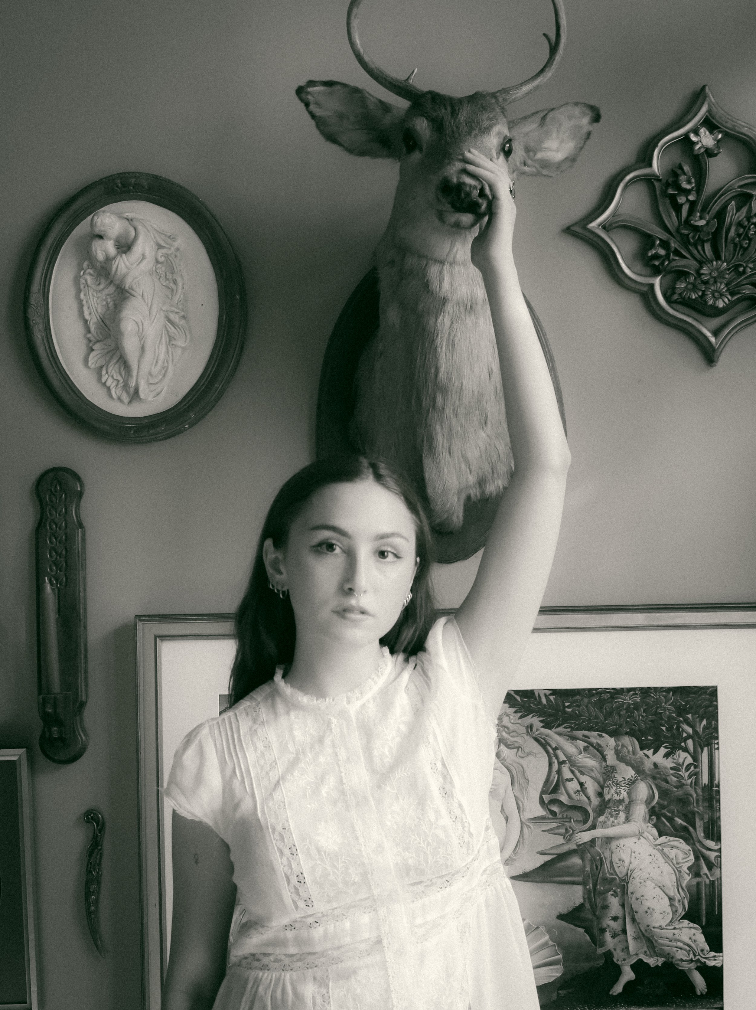 A young woman with dark hair and earrings, wearing a white dress, stands in front of a wall decorated with various framed artworks. She raises her left arm, with her hand covering her face, and a mounted deer head and decorative wall art are visible 