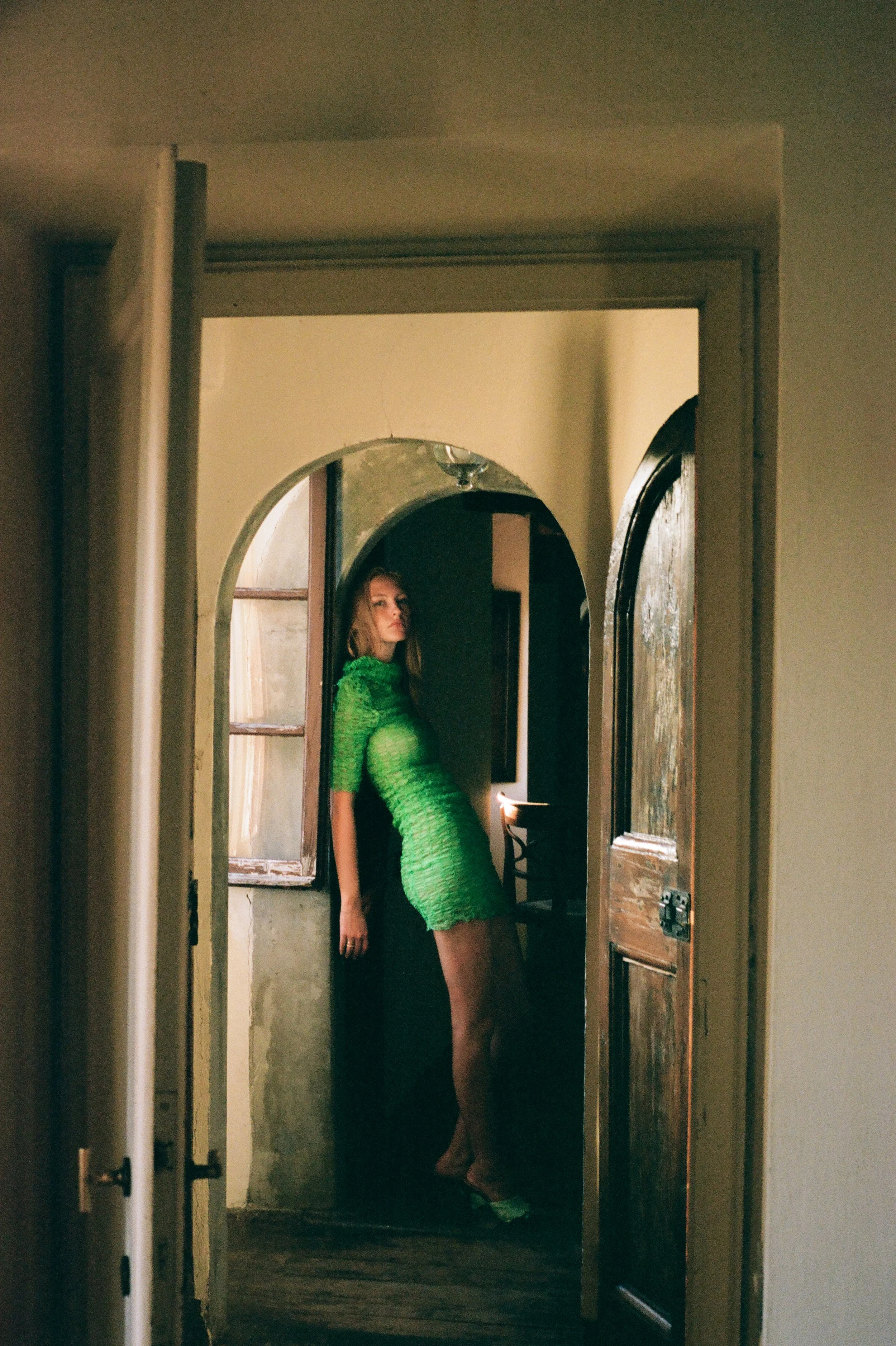 A woman in a bright green textured dress standing in front of a mirror in an indoor setting with wooden floors and a doorframe.