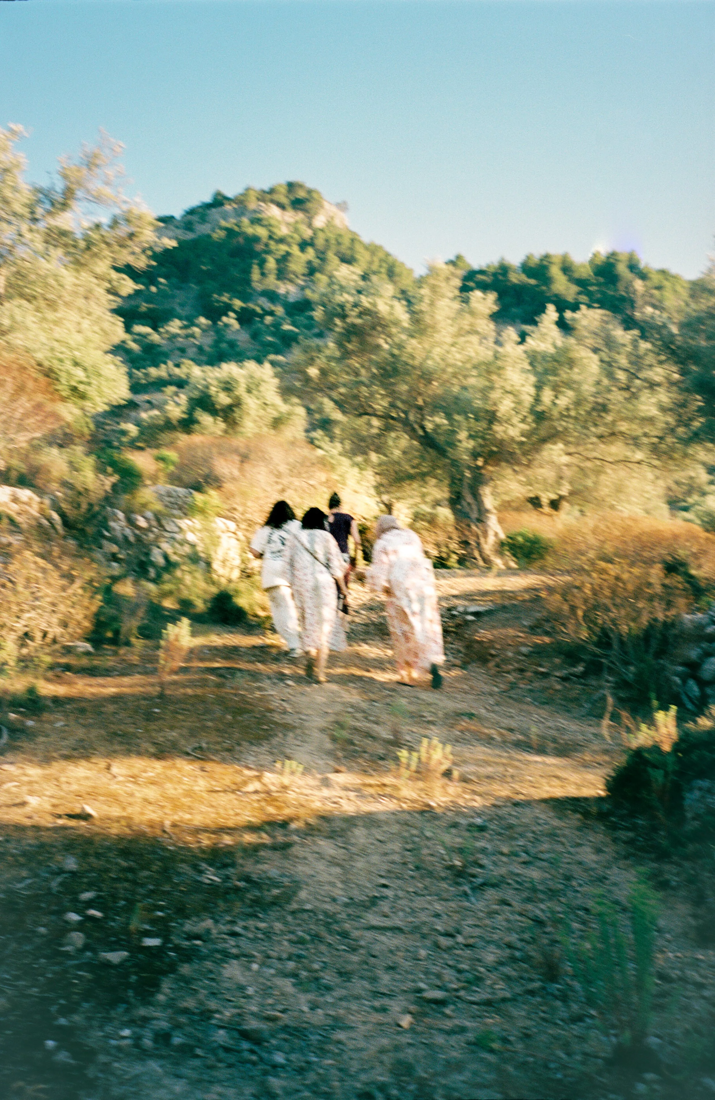 Four women dressed in pajamas walking together on a dirt path surrounded by trees and bushes in a natural outdoor setting.