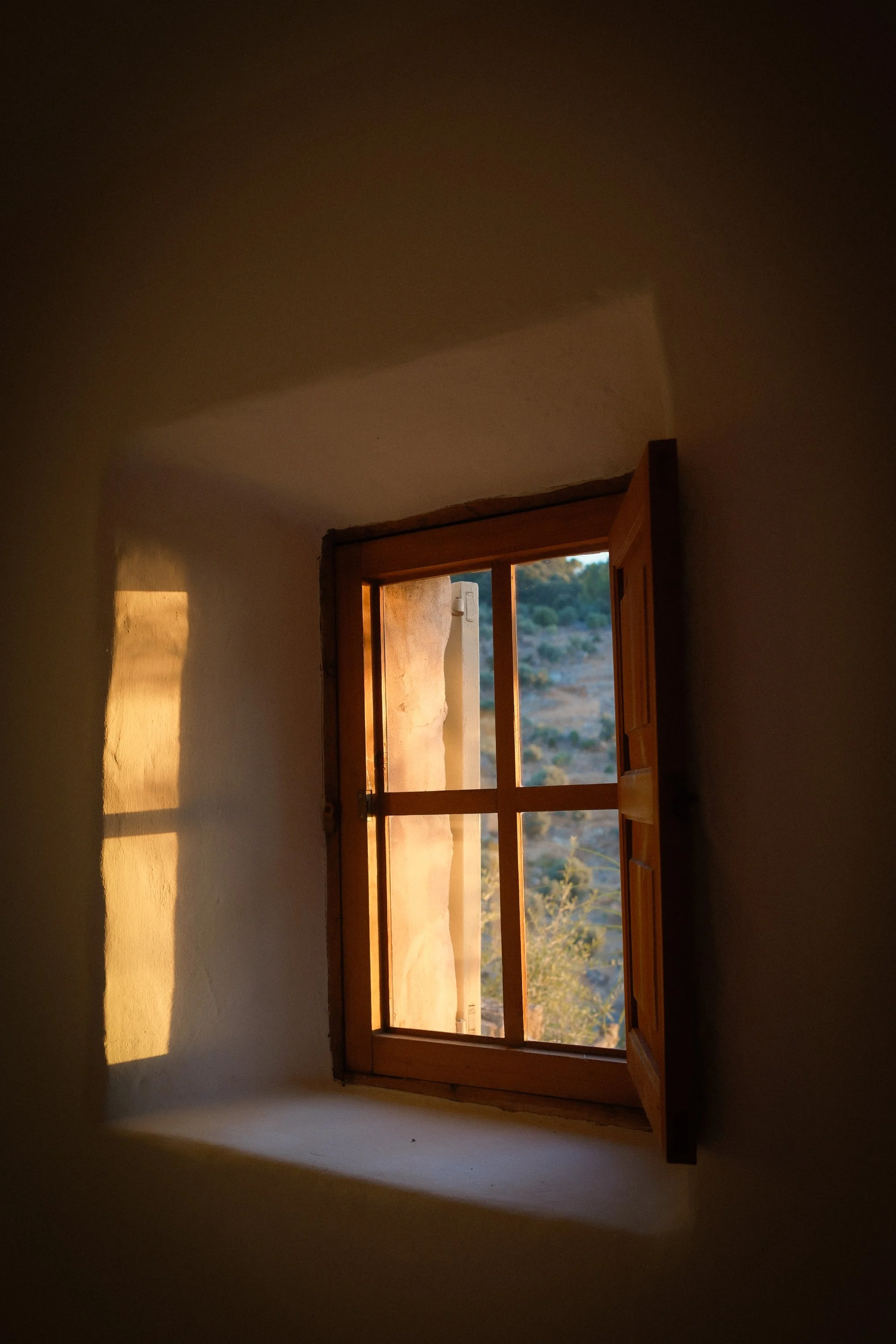 Sunlight coming through a wooden window in a plain white room, with a view of a distant landscape with hills and trees.