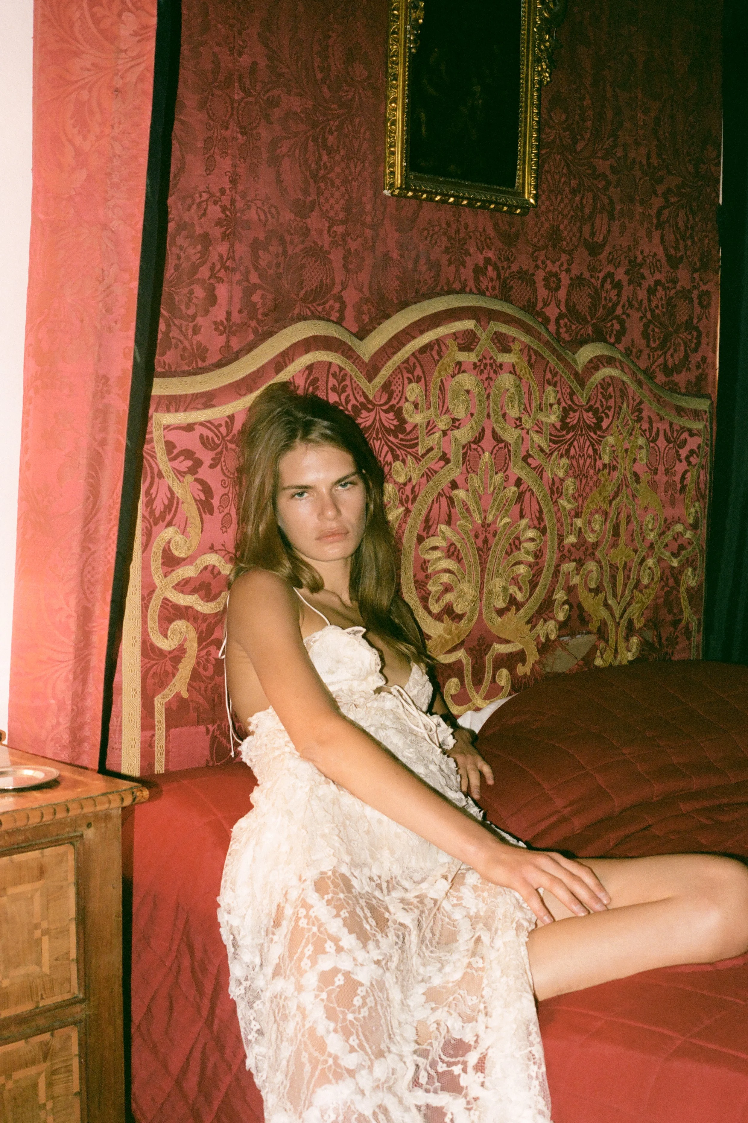 A young woman sitting on a bed in a richly decorated room, wearing a white lace dress, with a detailed red and gold headboard, ornate wallpaper, and framed artwork on the wall.