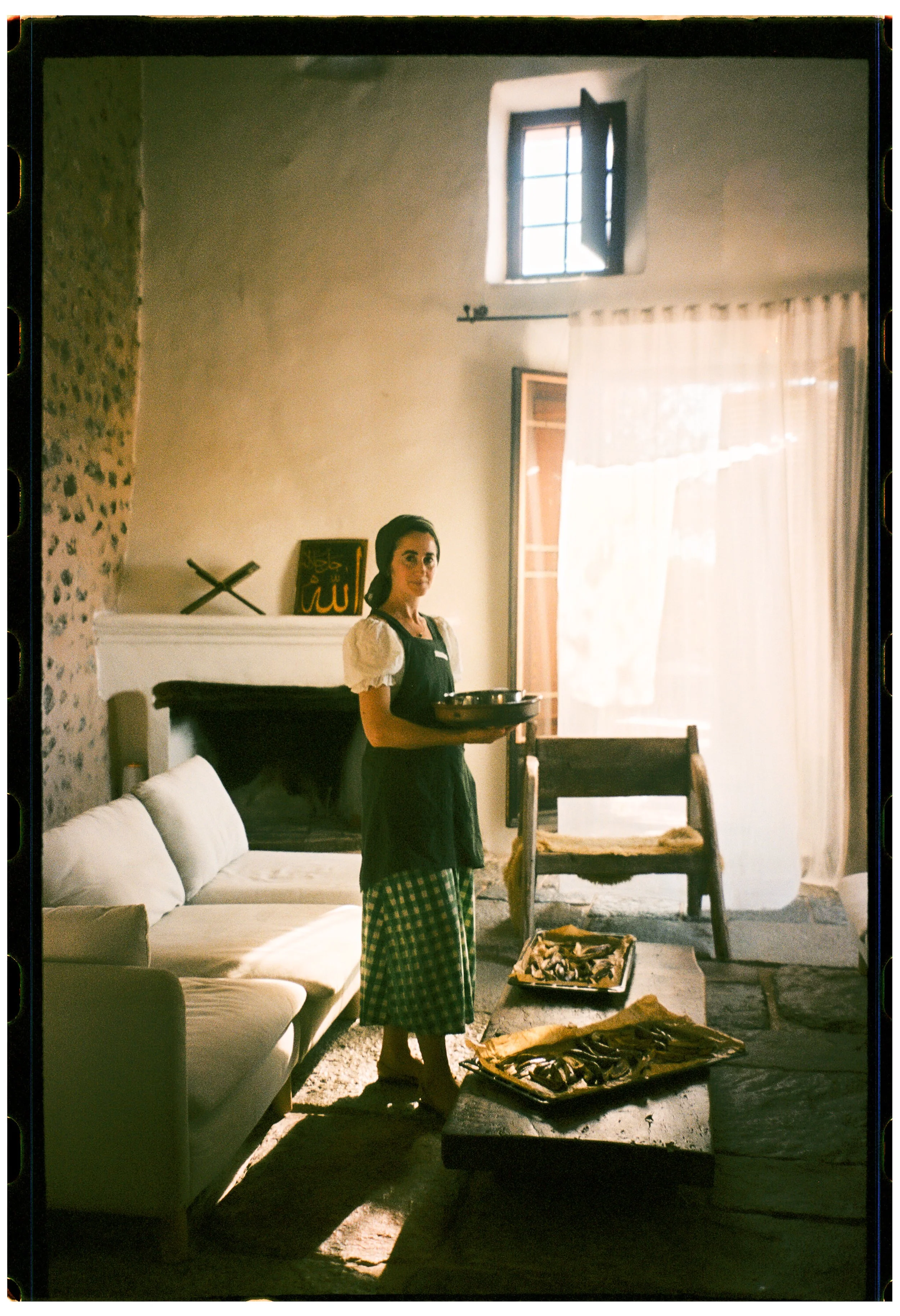 A woman standing in a room holding a tray. The room has sunlight coming through a window, a white sofa, a stone wall on the left, and a table with various objects on it.