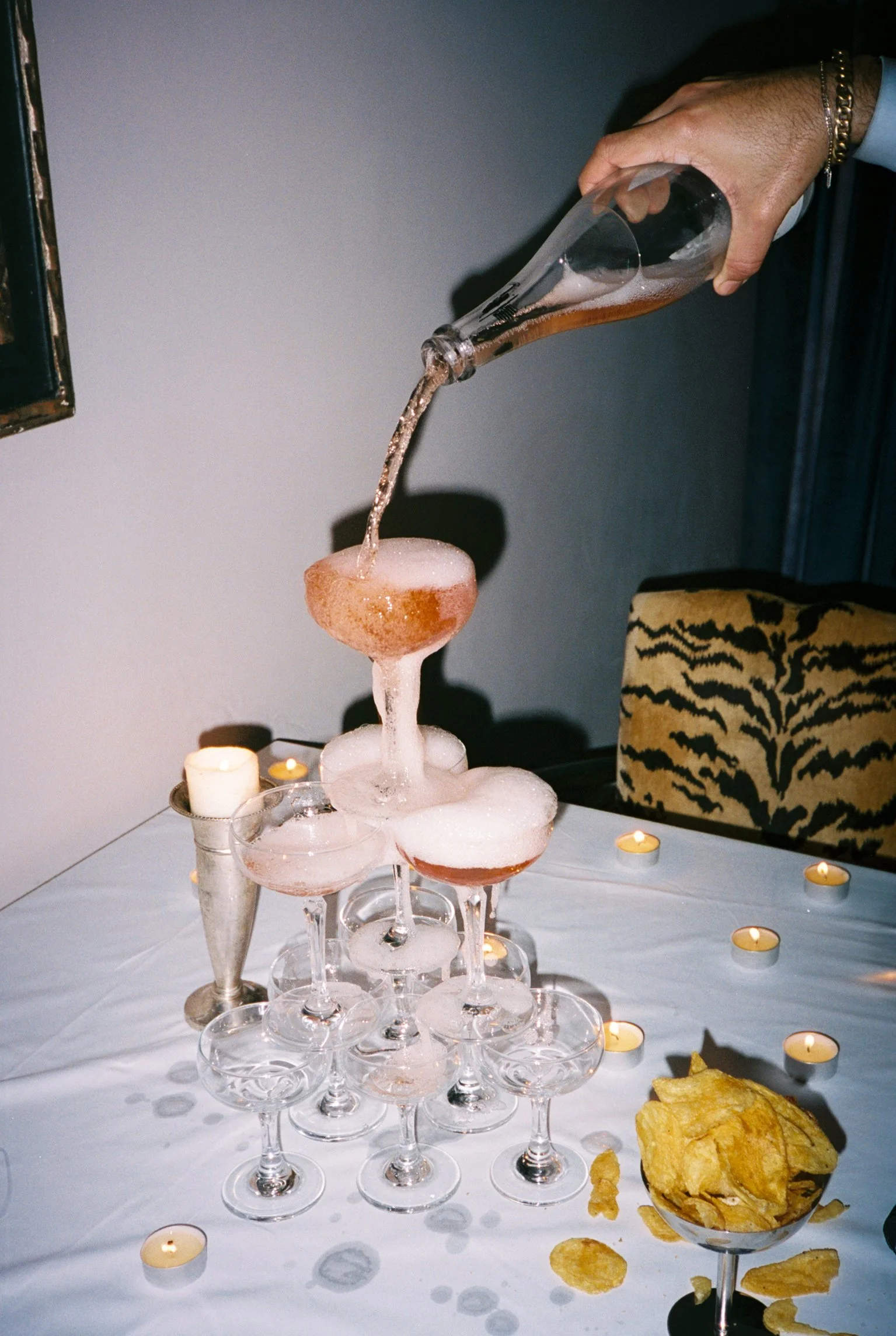A pyramid of empty champagne glasses on a table with a bottle pouring liquid into the top glass and surrounded by candles and snacks.