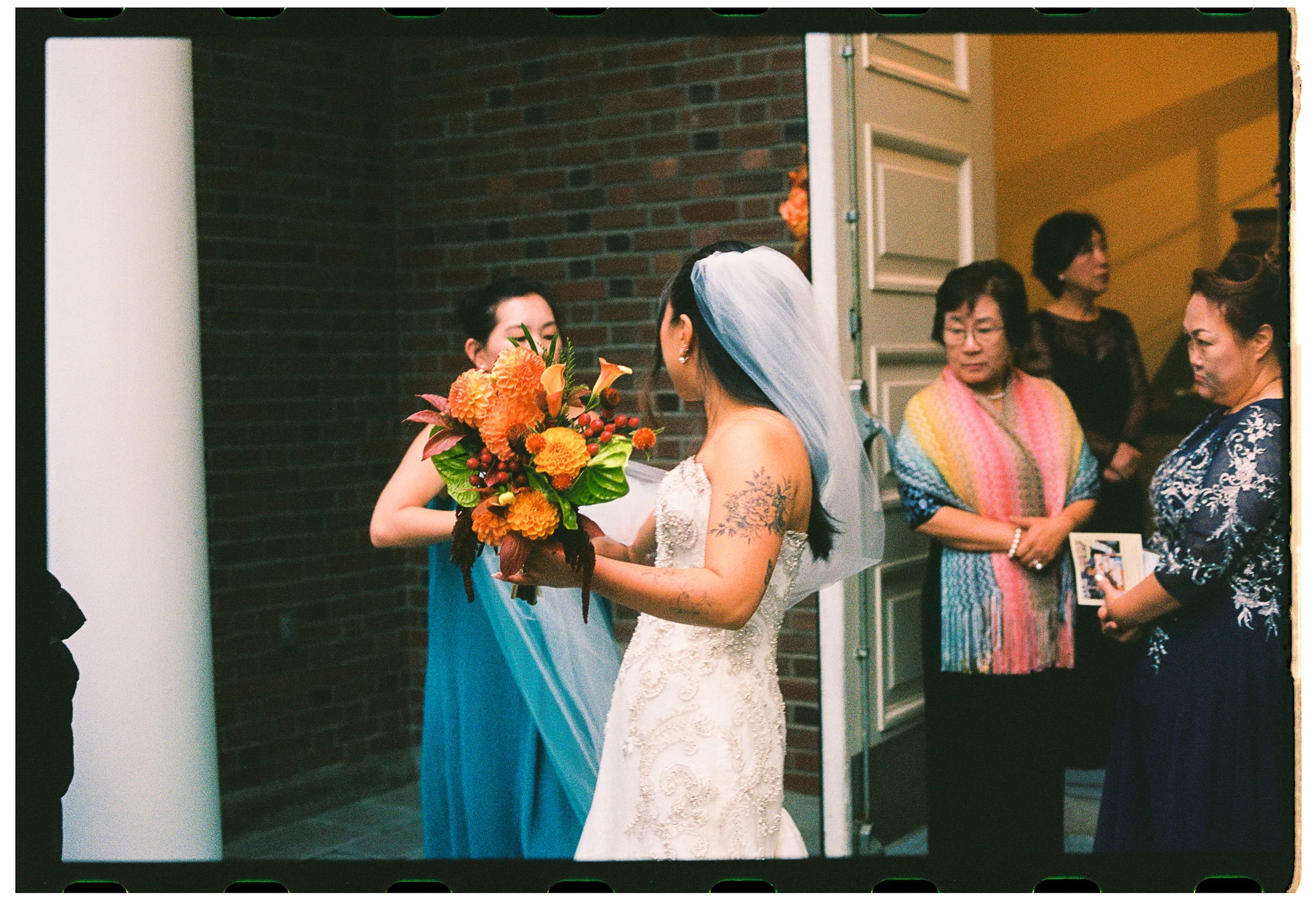 A bride with long silver hair and tattoos on her arm, wearing a white lace wedding dress, is receiving a bouquet of orange flowers from another woman at her wedding. Several guests, mostly women, are watching in the background.