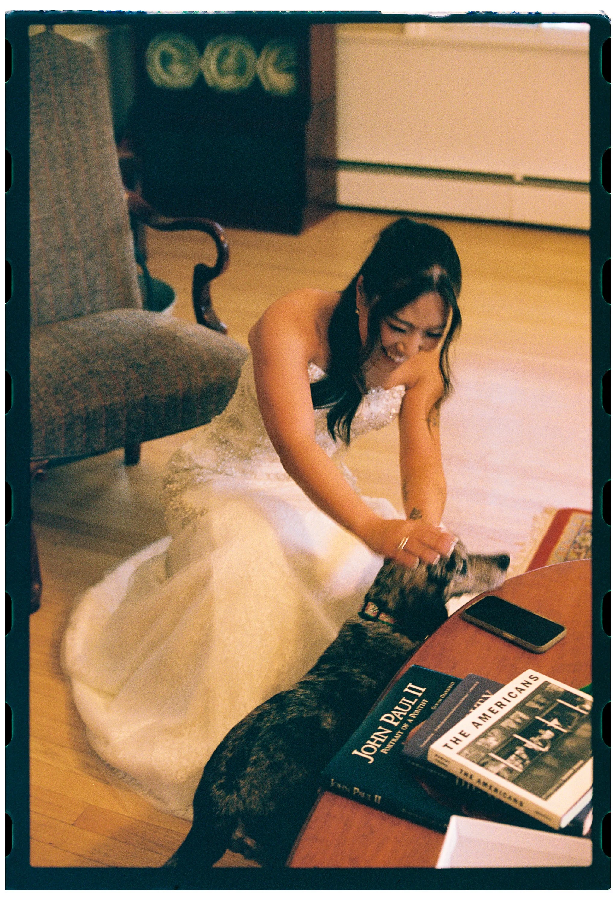 A woman in a wedding dress smiling and reaching down to pet a dog in a living room with wooden floors, a cushioned armchair, and a coffee table with books and a smartphone.